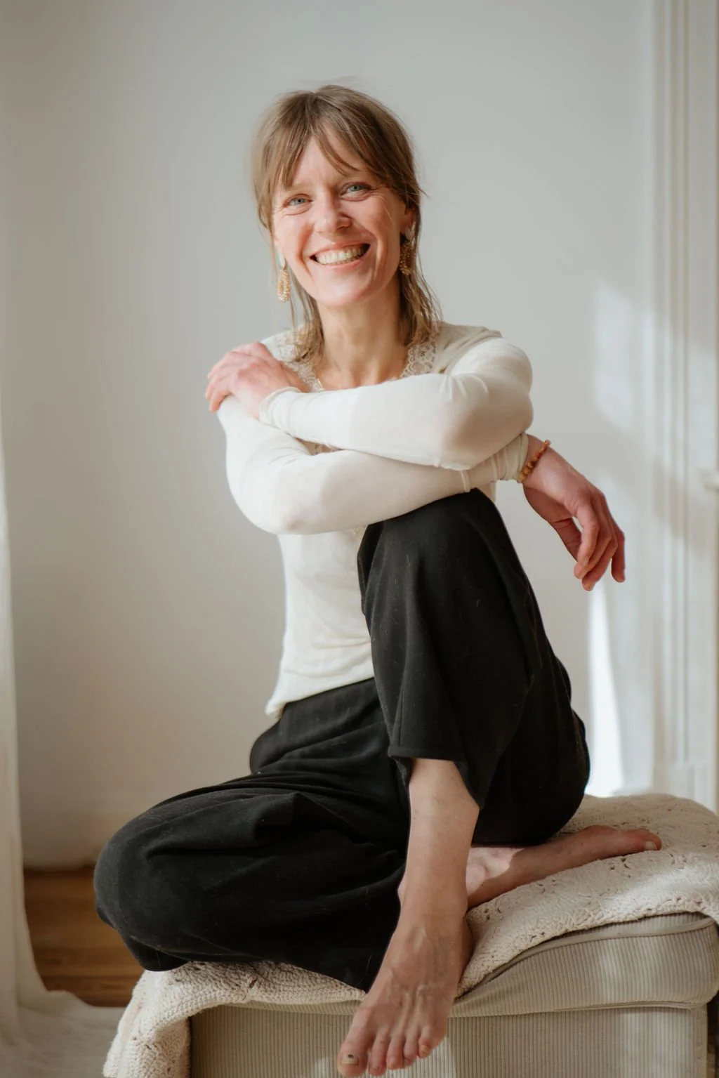 Eine lächelnde Frau sitzt auf einem beigefarbenen Sitz mit einem weißen Pullover und schwarzen Hosen, die Beine angezogen, vor einem hellen Hintergrund.