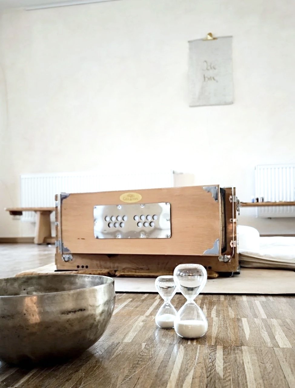 Ein Holzkasten mit Metallplatten liegt auf dem Boden, im Vordergrund sind eine Metallschale und zwei Sanduhren auf einem Holztisch.