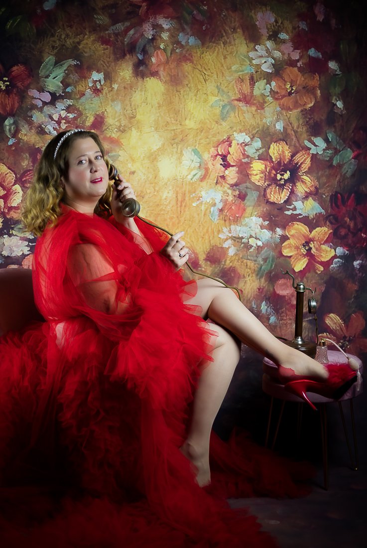 Femme en robe rouge, assise sur un canapé, tenant un téléphone ancien au visage, devant un mur avec un motif floral coloré.