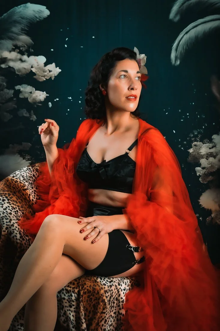 Femme assise portant un ensemble noir et une robe rouge en satin, assise sur un fauteuil à motif léopard, avec un fond artistique représentant des nuages et un ciel sombre.