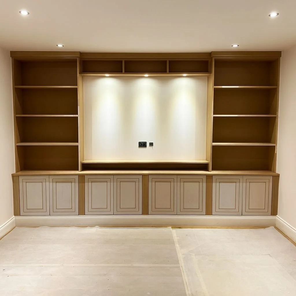 Empty wooden built-in bookshelves with lighting in a room with beige walls and floor.  Carpentry, construction and Renovation by DA Build in London, Surrey and Greater London.