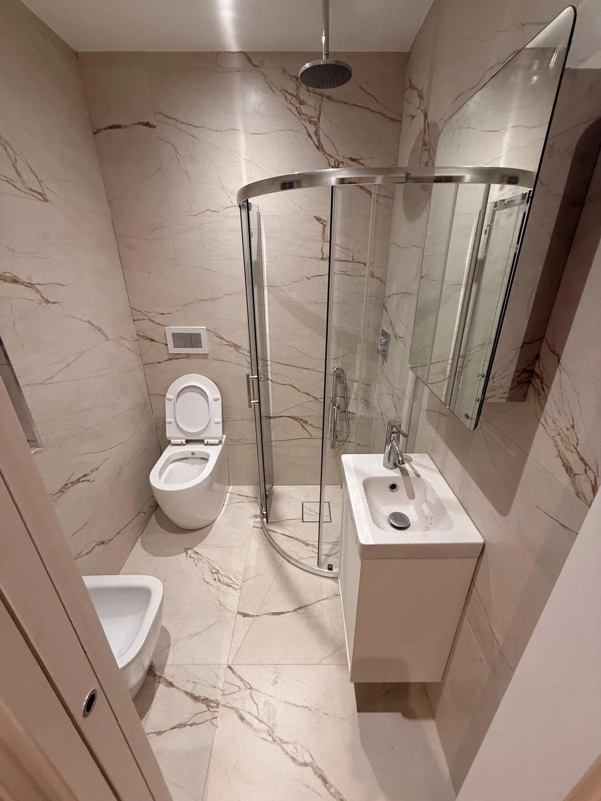 Small modern bathroom with marble walls and floor, glass shower enclosure, wall-mounted sink, mirror, and toilet. Bathroom renovations.  Carpentry, construction and Renovation by DA Build in London, Surrey and Greater London.