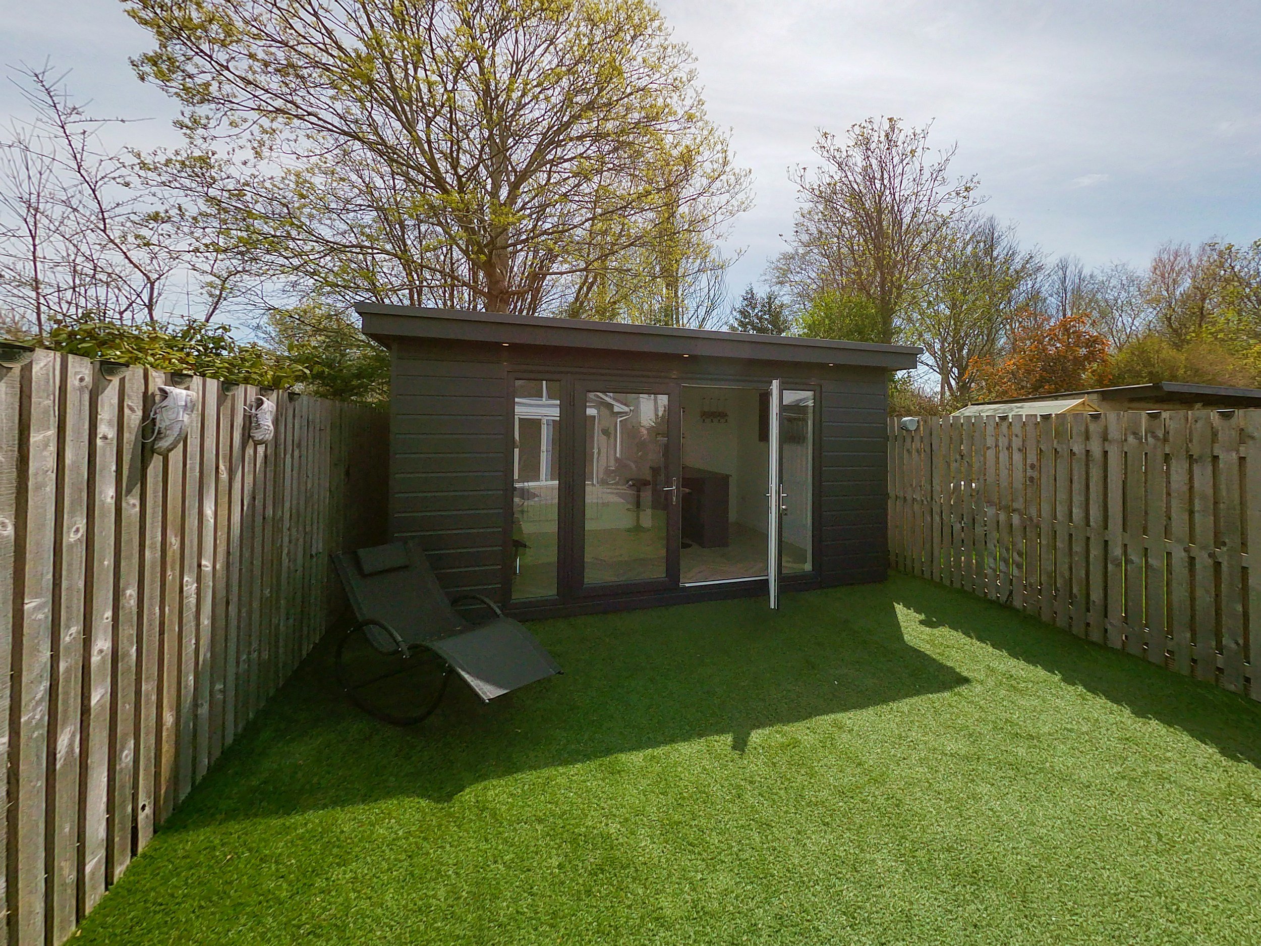 A backyard with a small black shed, a reclining lawn chair, a grassy lawn, and a wooden fence. There are trees in the background and a cloudy sky.