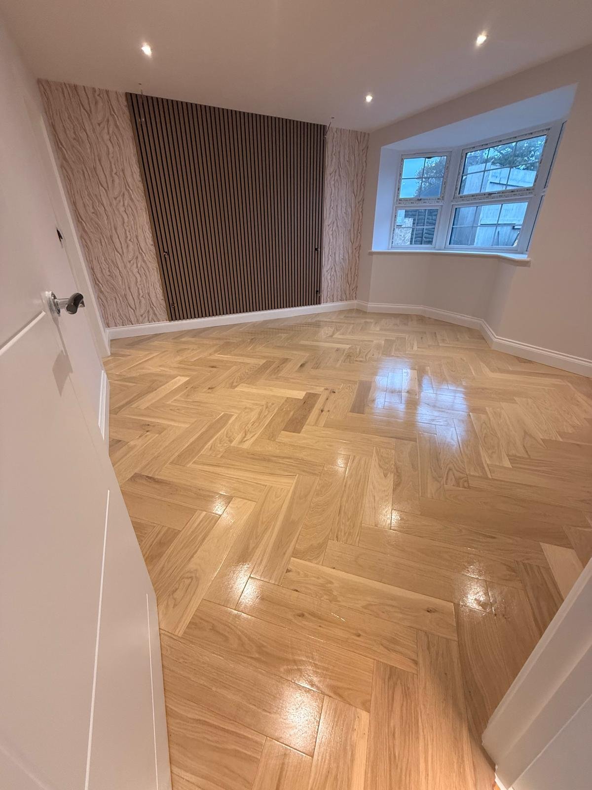 Polished wooden herringbone floor, white walls, a window with a view of a garden, and wooden slats on the wall.  Carpentry, construction and Renovation by DA Build in London, Surrey and Greater London.