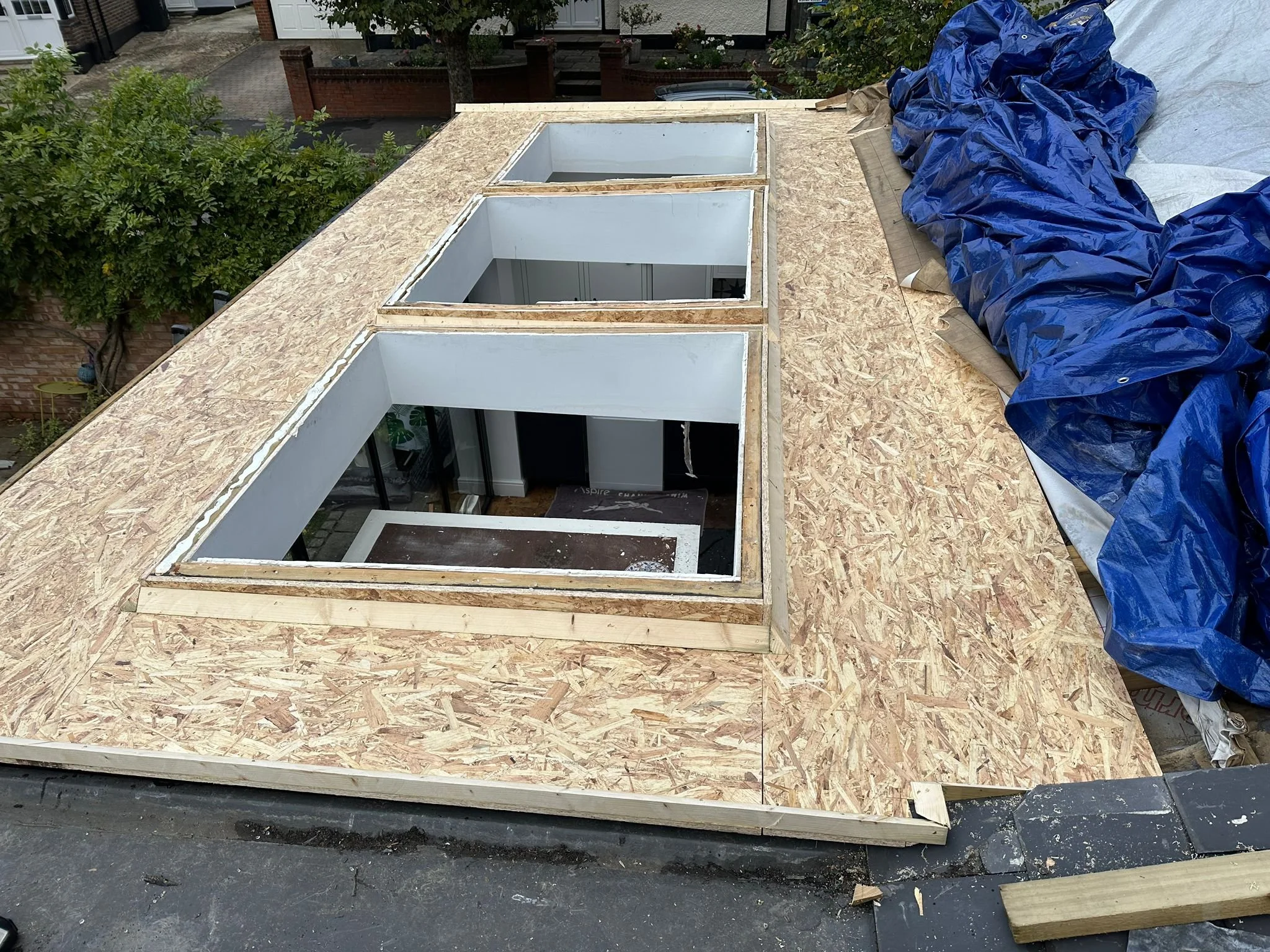 Construction of a new flat roof on a building, with three large skylights installed, and blue tarps covering some materials on the right side. Extneions, sky lights, Carpentry and Renovation by DA Build in London, Surrey and Greater London.