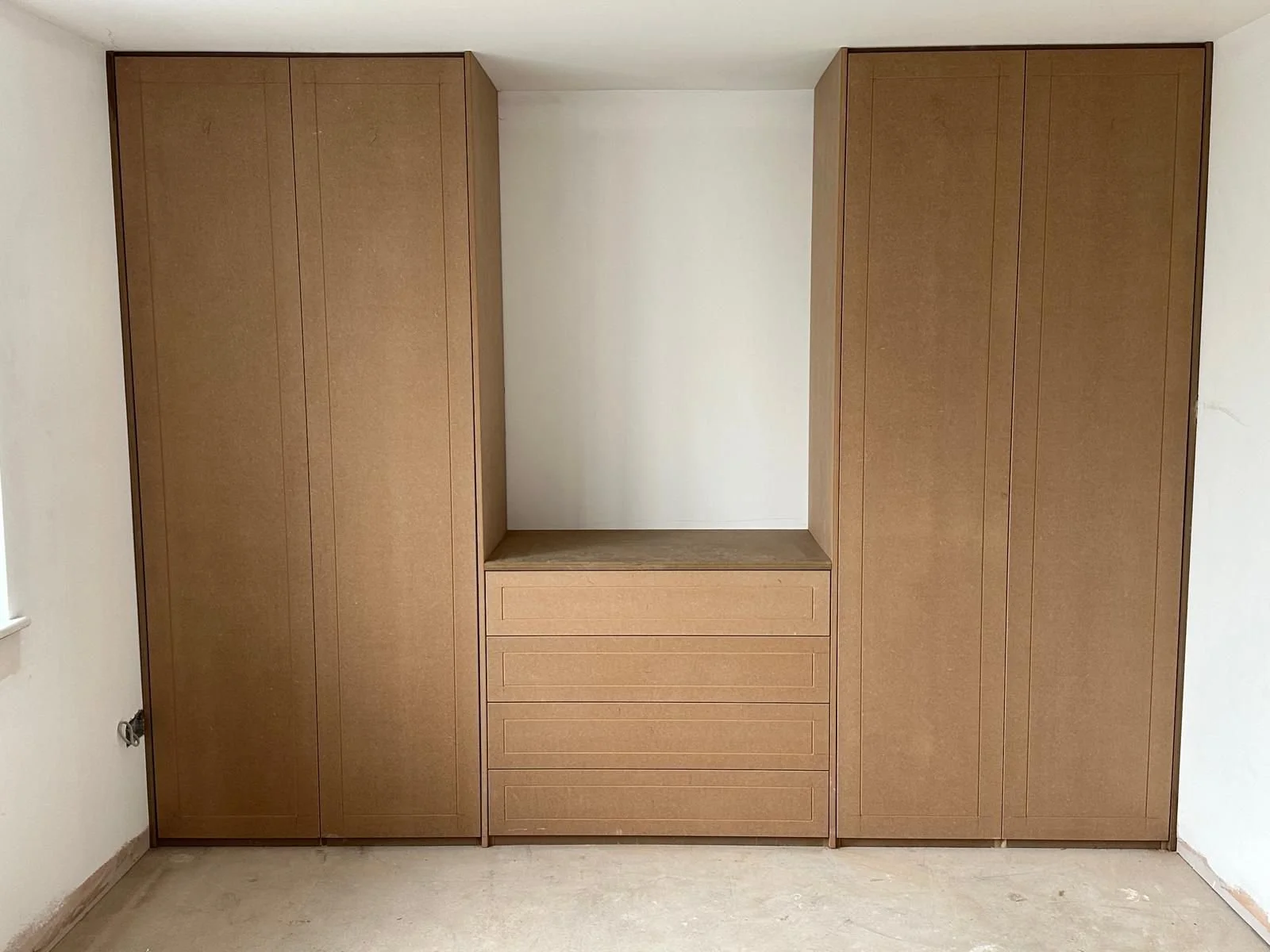Built-in wooden wardrobe with four large compartments and four drawers in the middle, in a room with white walls and a concrete floor.  Carpentry, construction and Renovation by DA Build in London, Surrey and Greater London.