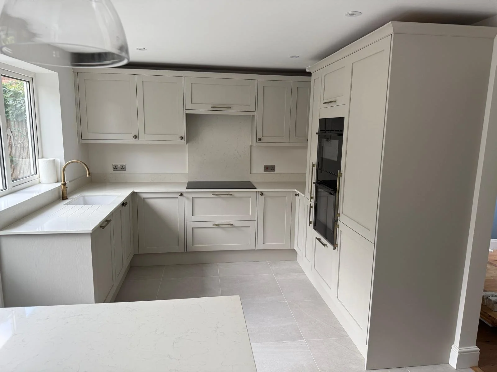 Modern white kitchen with upper and lower cabinets, a central electric cooktop, built-in double oven, a window with a gold faucet sink, and a light-colored tile floor.  Carpentry, construction and Renovation by DA Build in London, Surrey and Greater 