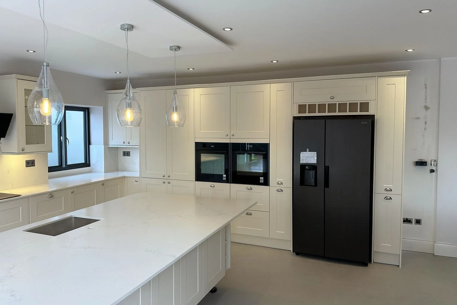 Modern kitchen with white cabinets, marble island, black refrigerator, built-in ovens, and hanging light fixtures.  Carpentry, construction and Renovation by DA Build in London, Surrey and Greater London.