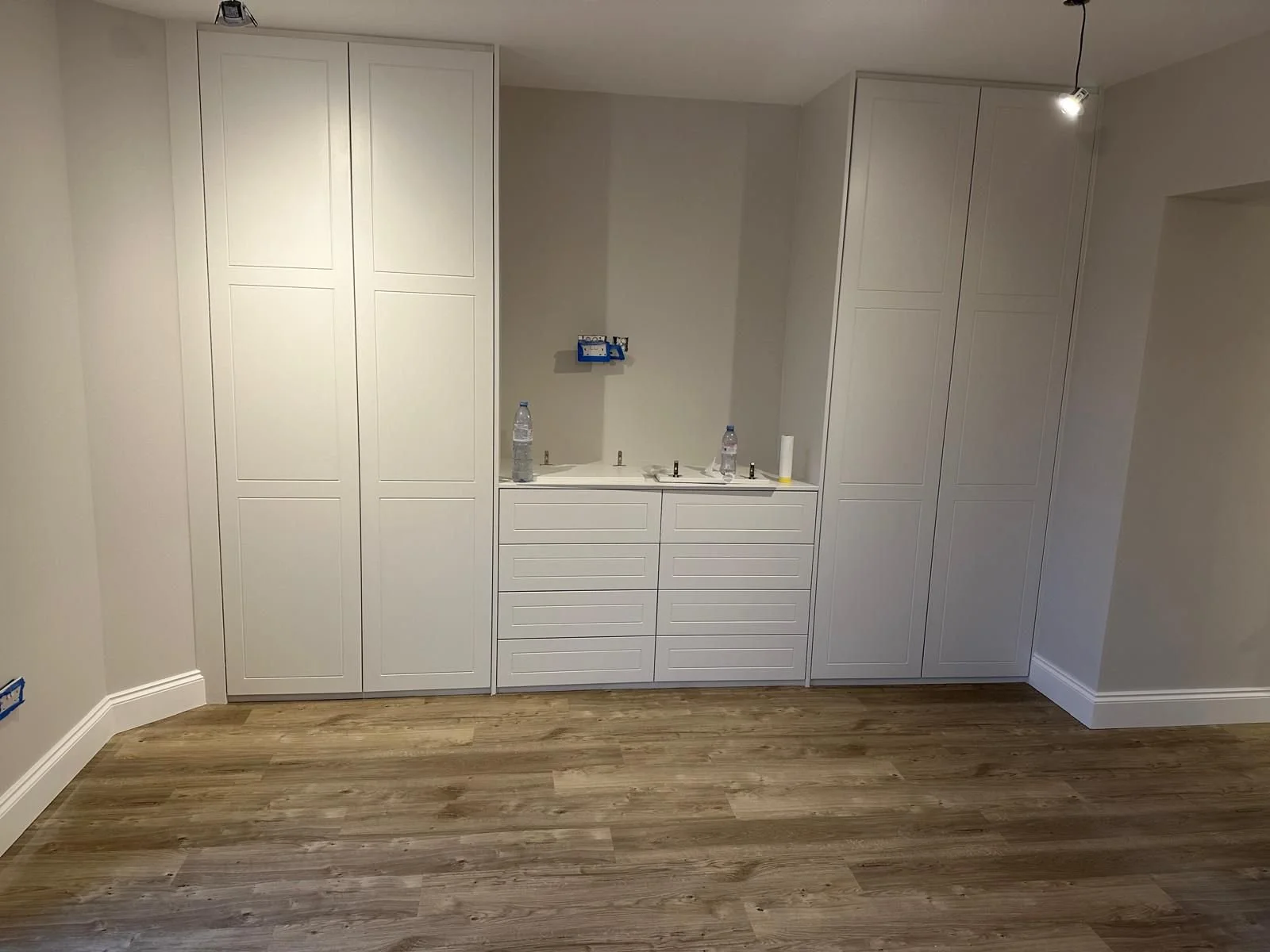 Room with white built-in cabinets and drawers, with electrical outlets and two water bottles on the countertop, on a wood floor.  Carpentry, construction and Renovation by DA Build in London, Surrey and Greater London.