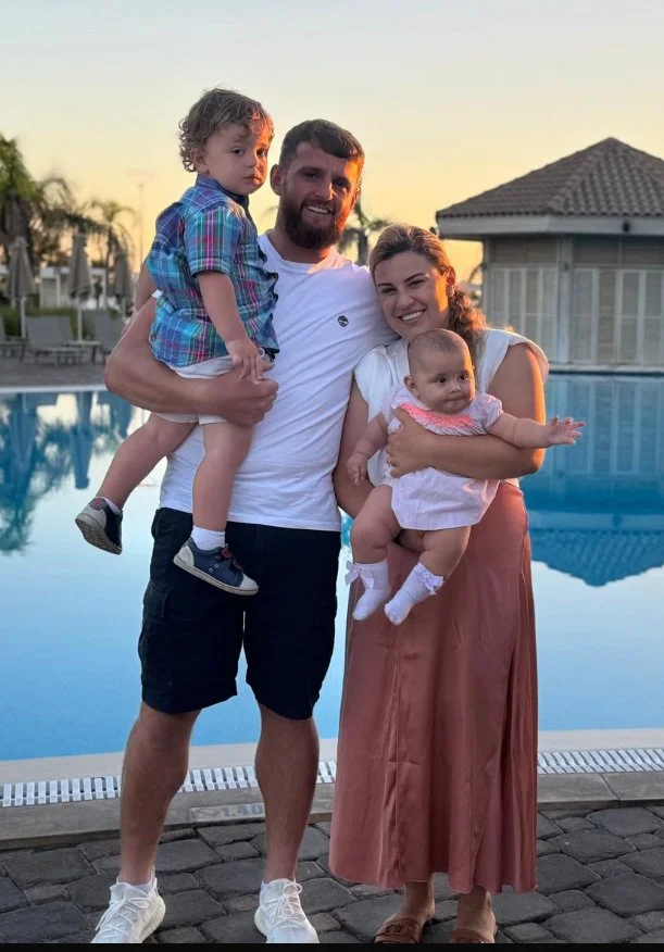 A family of four standing by a swimming pool at sunset. The father is holding a young boy on his shoulder, and the mother is holding a baby girl in her arms. All are smiling.