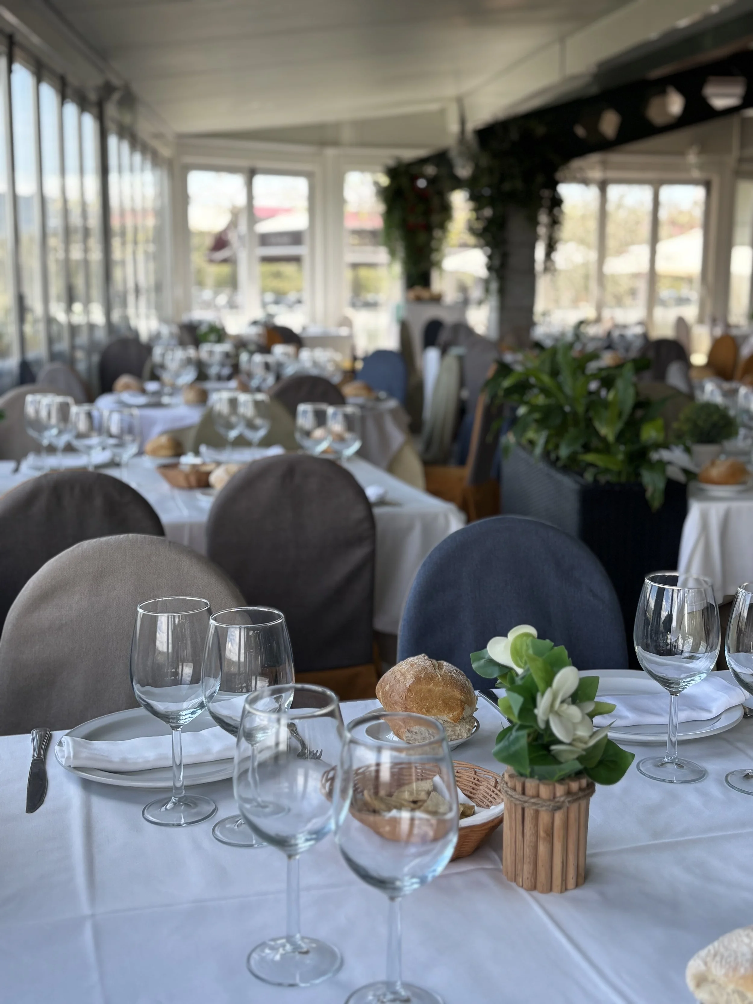 Mesa elegante en un restaurante con platos, vasos y pan, decorada con flores y plantas verdes, en un ambiente bien iluminado con ventanas grandes.