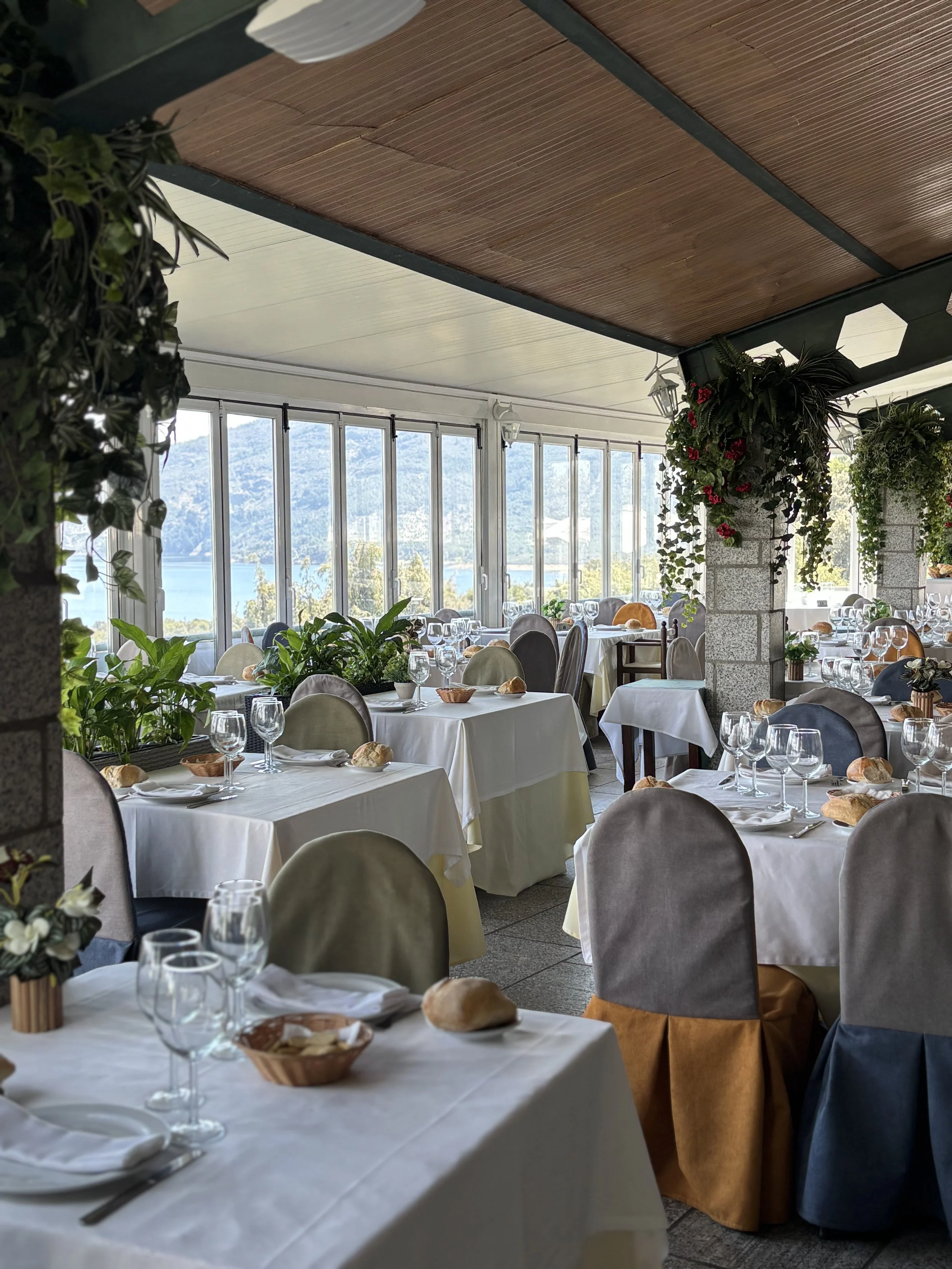 Restaurante con mesas vestidas con manteles blancos, decoradas con panes y cristalería, rodeado de plantas y con ventanas con vista a un paisaje natural.