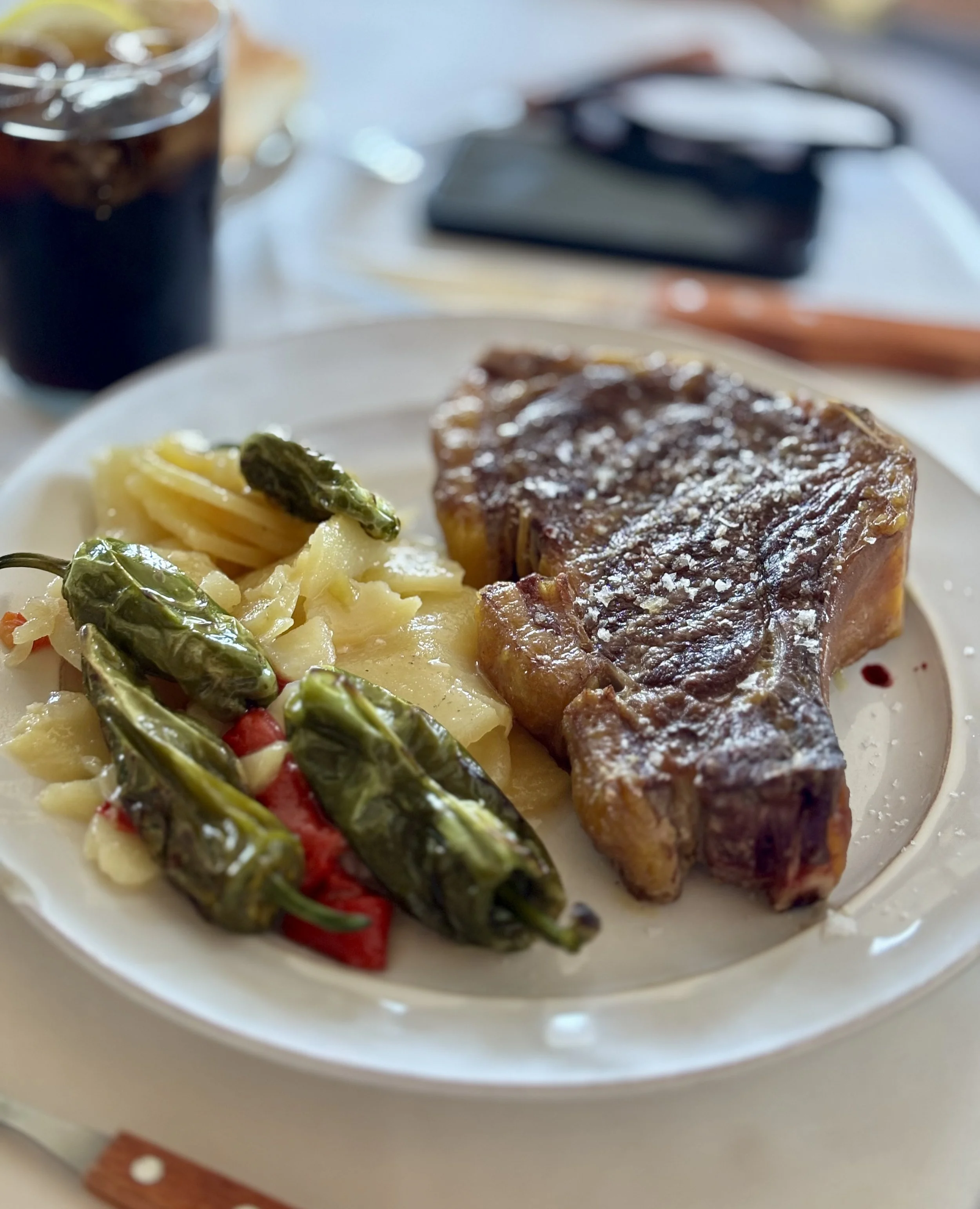 Plato con carne a la parrilla, papas cocidas con pimientos verdes y rojos, y una bebida oscura. En el fondo hay un teléfono y unos cubiertos.