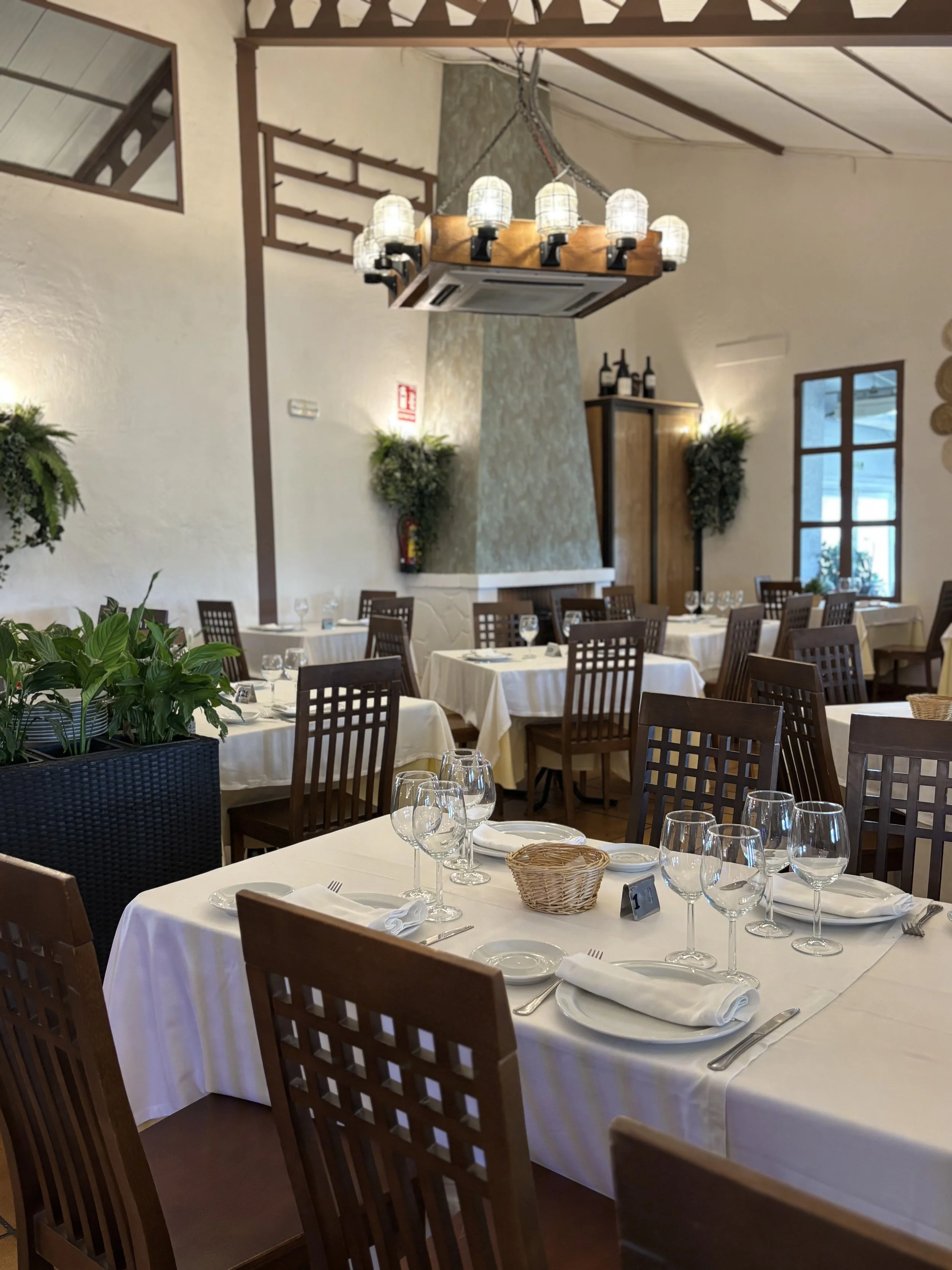 Comedor elegante con mesas cubiertas con manteles blancos, cristalería, vajilla y cubiertos, elegante ambiente decorado con plantas verdes y lámpara de techo con varias luces, ventanas que dejan entrar luz natural y paredes de color beige.