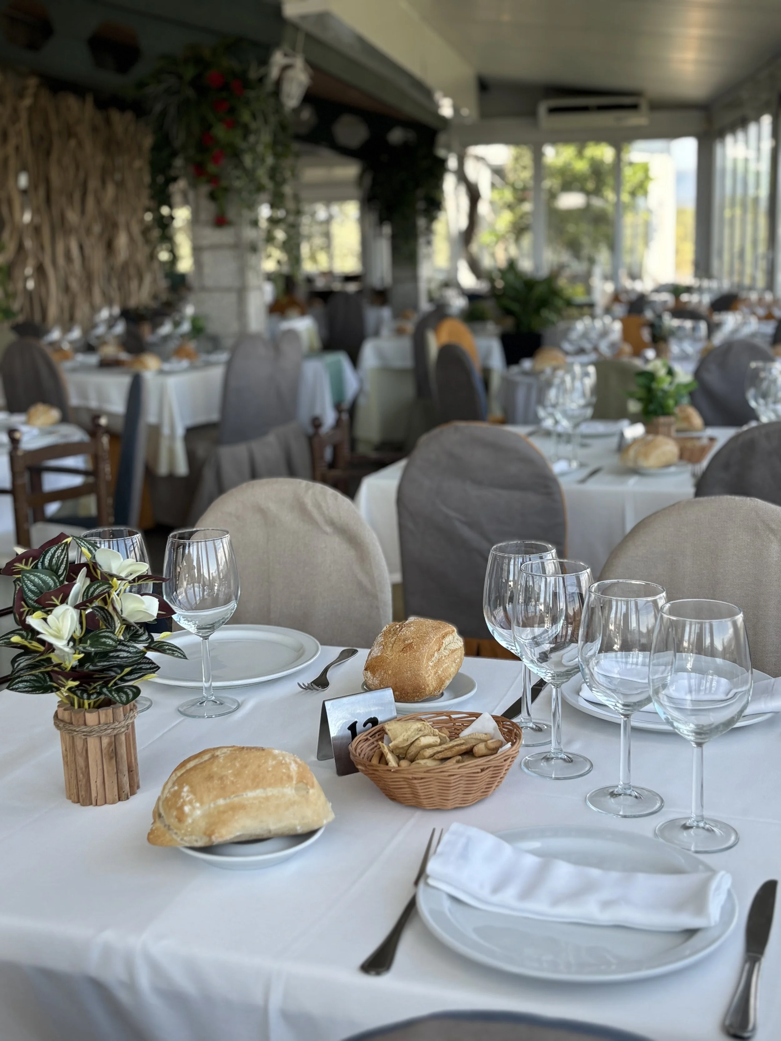 Mesa elegant preparada para desayuno o comida en un restaurante, con vasos de agua, pan, flores decorativas y cubiertos, en un ambiente con muchas ventanas y luz natural.