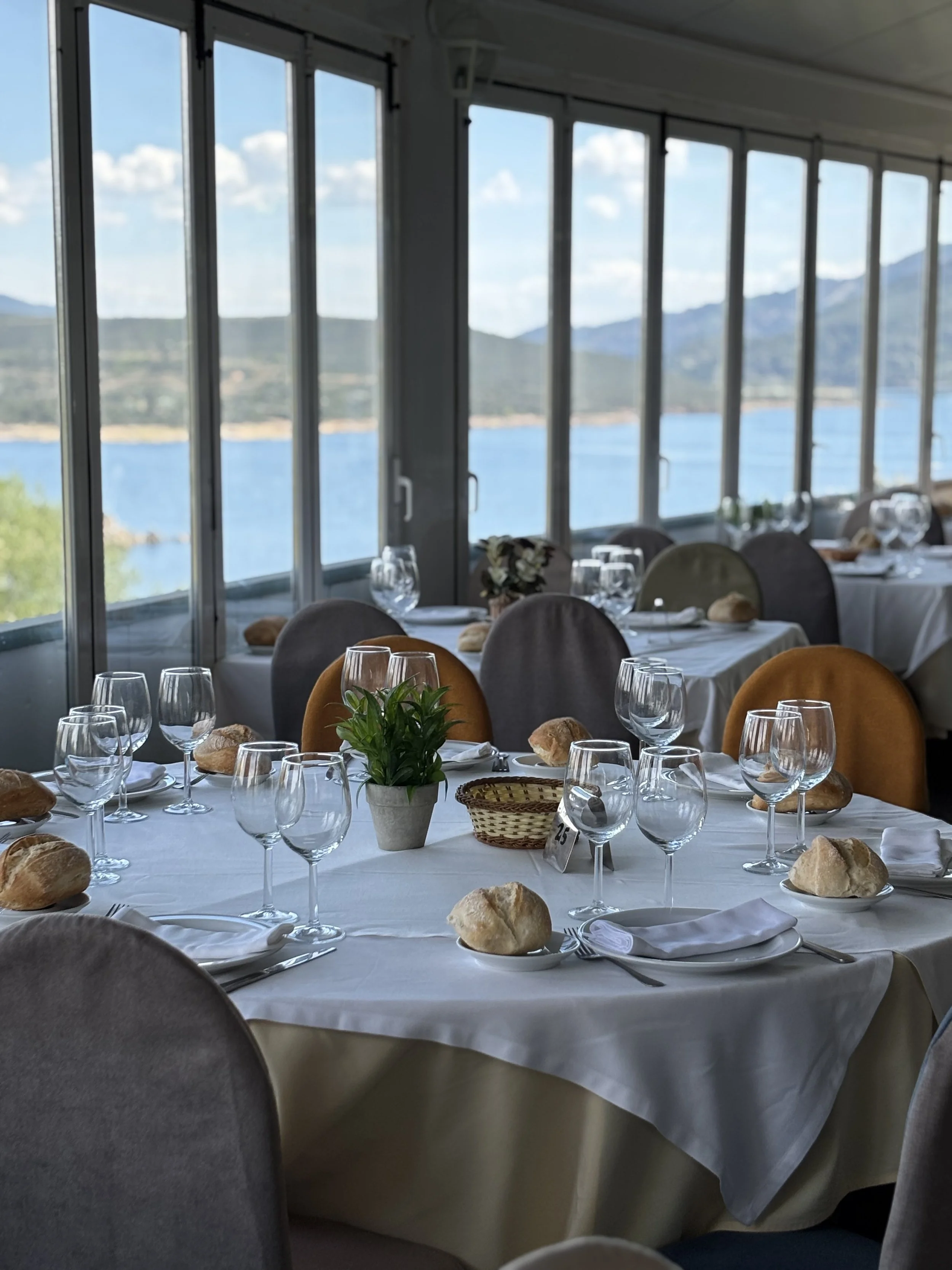 Interiores de un restaurante con mesas vestidas con manteles blancos, copas de vino, pan y accesorios, junto a ventanas grandes con vista a un paisaje montañoso y un cuerpo de agua.
