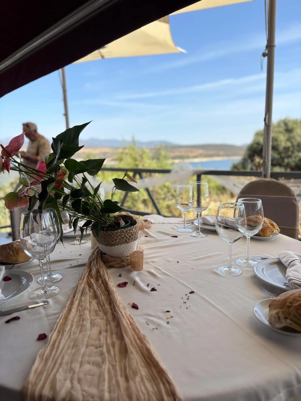 Mesa decorada con un mantel claro, un centro de flores en una maceta de rafia, varias copas y platos, pan sobre platos, y algunos pétalos de rosa, con vista panorámica de un paisaje natural y cielo azul al fondo.