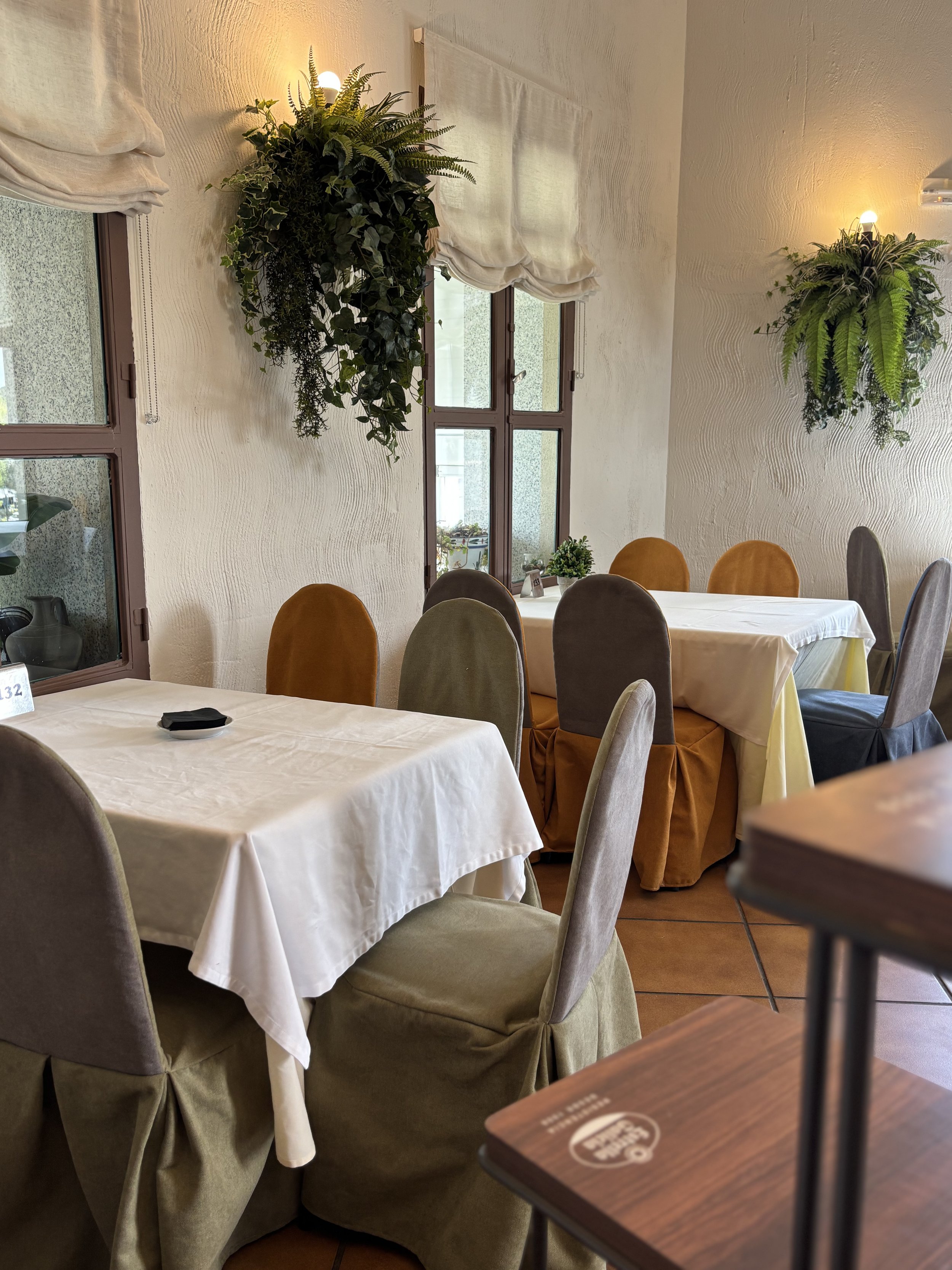 Vista de un comedor con mesas cubiertas con manteles blancos, sillas en tonos beige, y paredes decoradas con plantas y luces. Hay ventanas con cortinas blancas en el fondo.