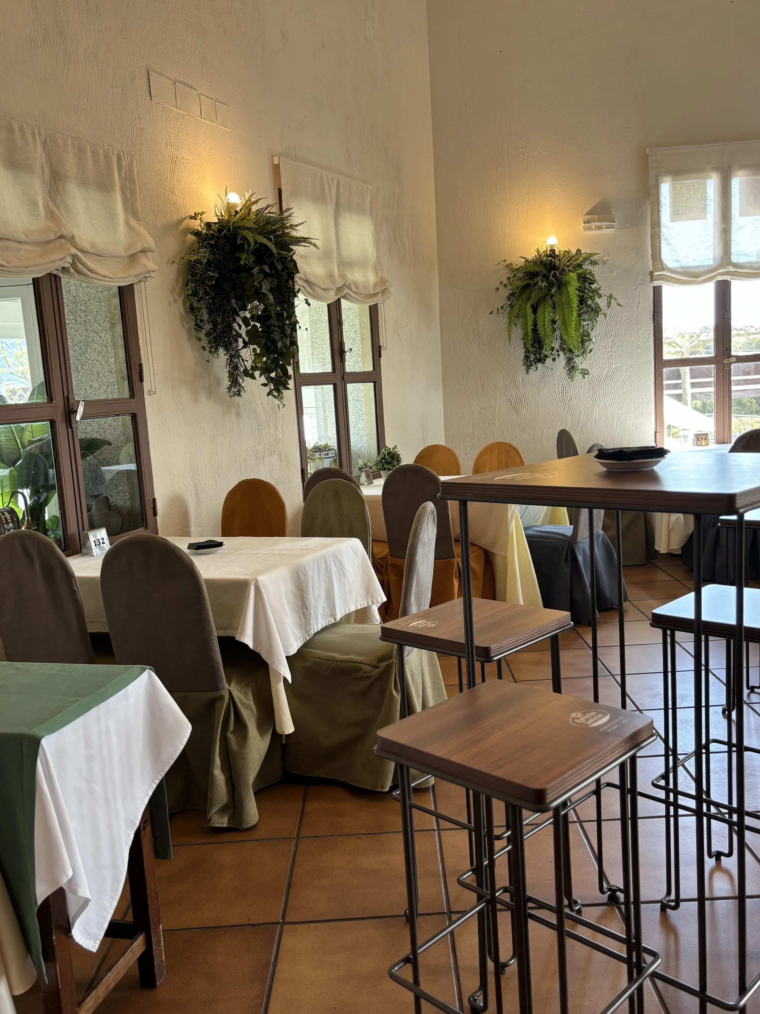 Interior de un restaurante con mesas con manteles blancos, sillas y paredes decoradas con plantas colgantes y cortinas. Hay ventanas que dejan entrar luz natural.