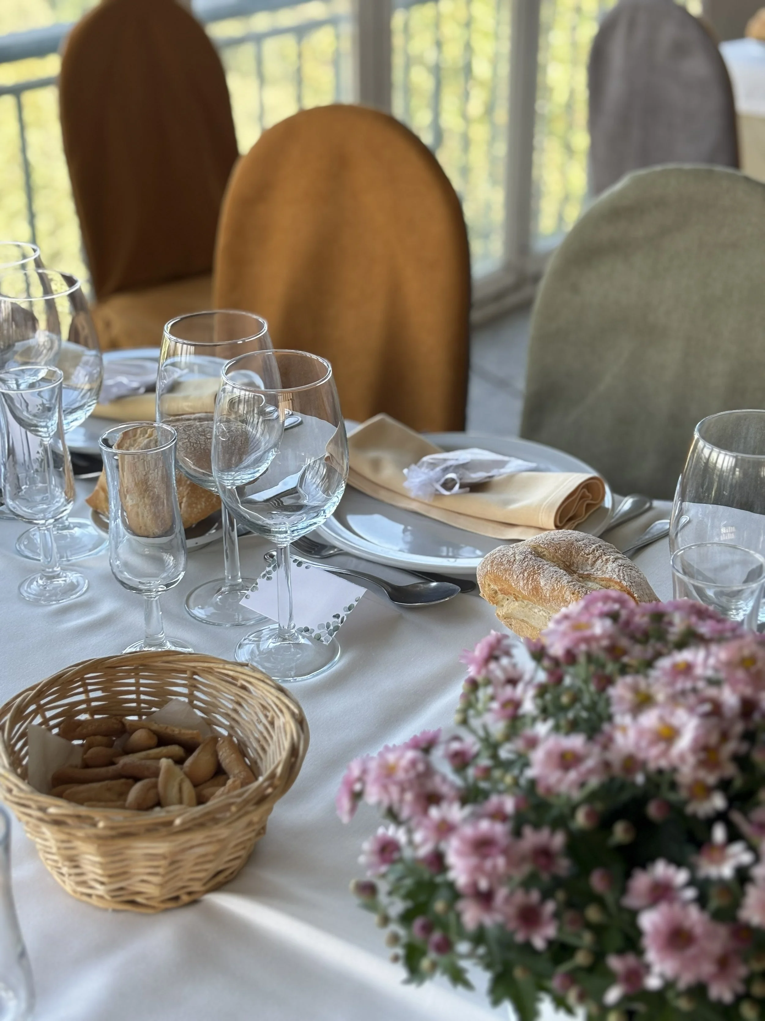 Mesa decorada para una comida o reunión, con copas, panes, un pequeño platillo con galletas o snacks, y un ramo de flores rosas en primer plano.