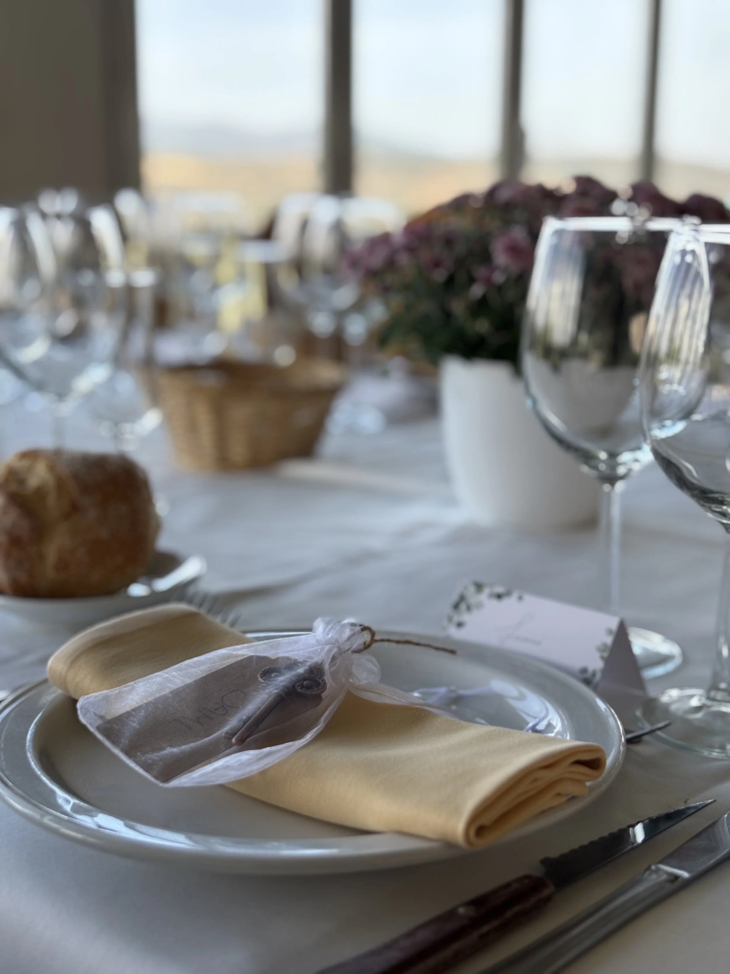 Una mesa decorada con copas de vino, platos con comida y vasos, en un entorno bien iluminado con ventanas grandes al fondo.