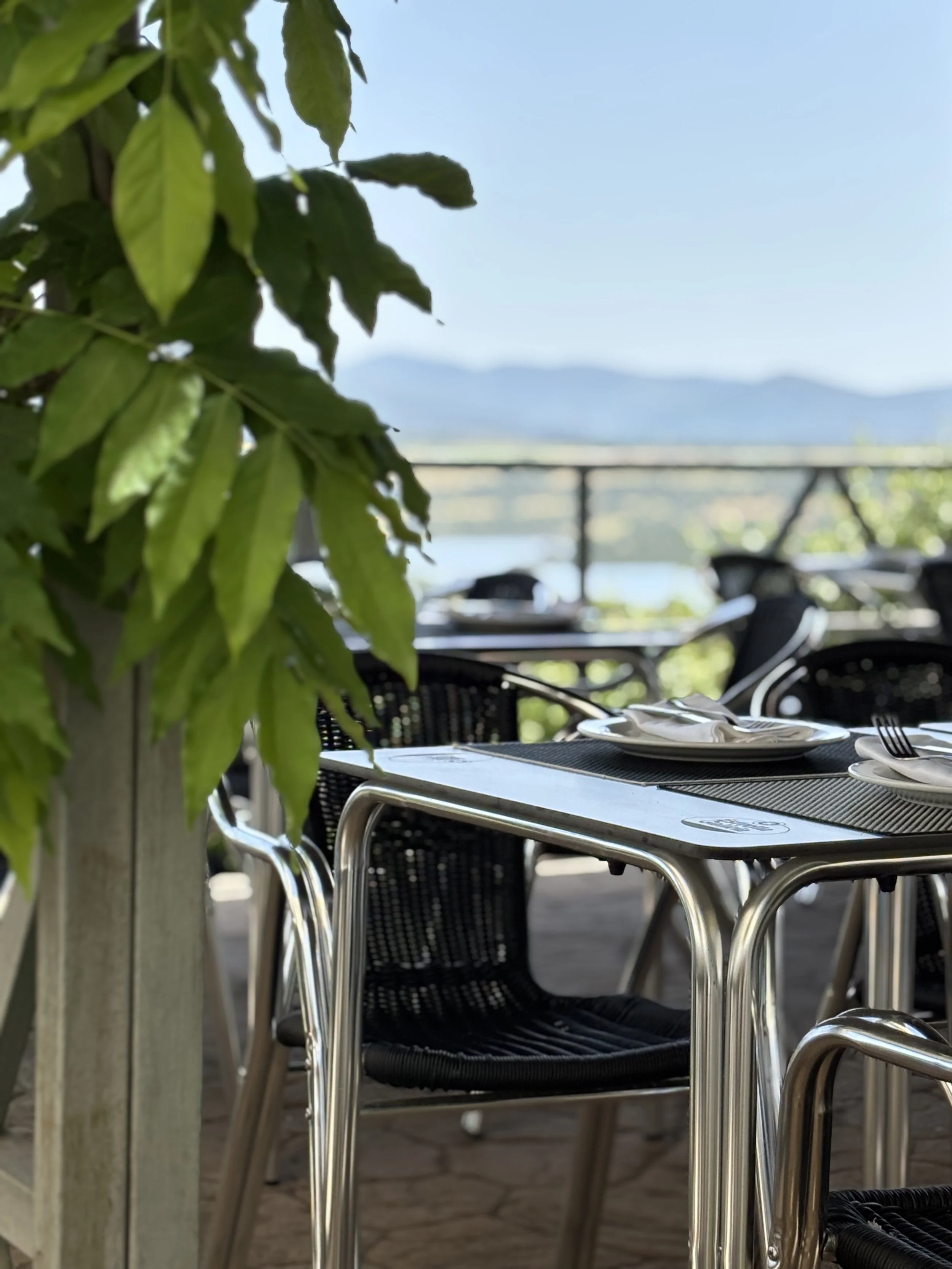 Mesas y sillas en un restaurante al aire libre con vista a un lago y montañas.