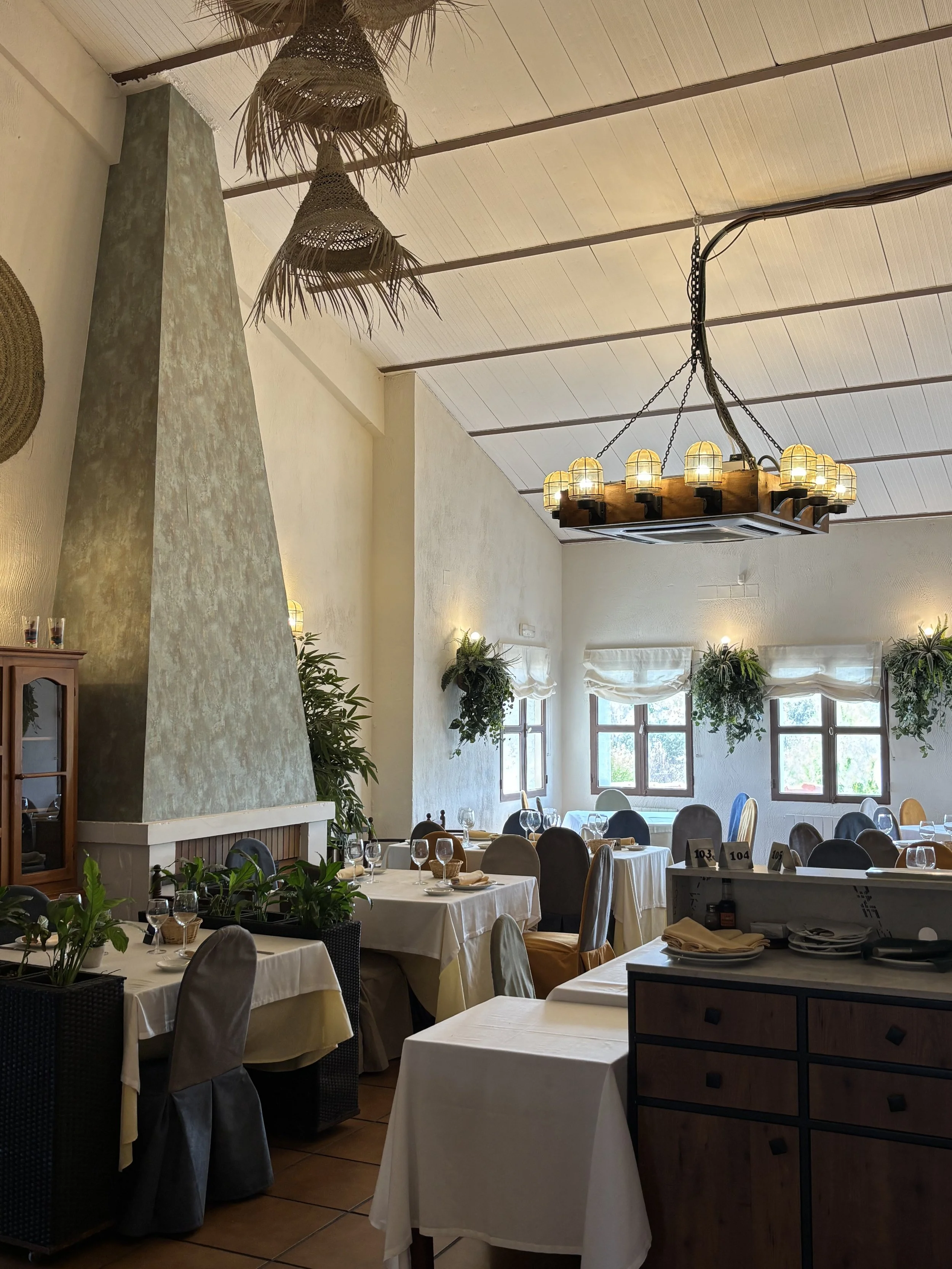 Comedor elegante con mesas vestidas de manteles blancos, copas y platos. Decoración con plantas y ventanas con cortinas. Iluminación cálida y lámparas de techo en estilo rústico.