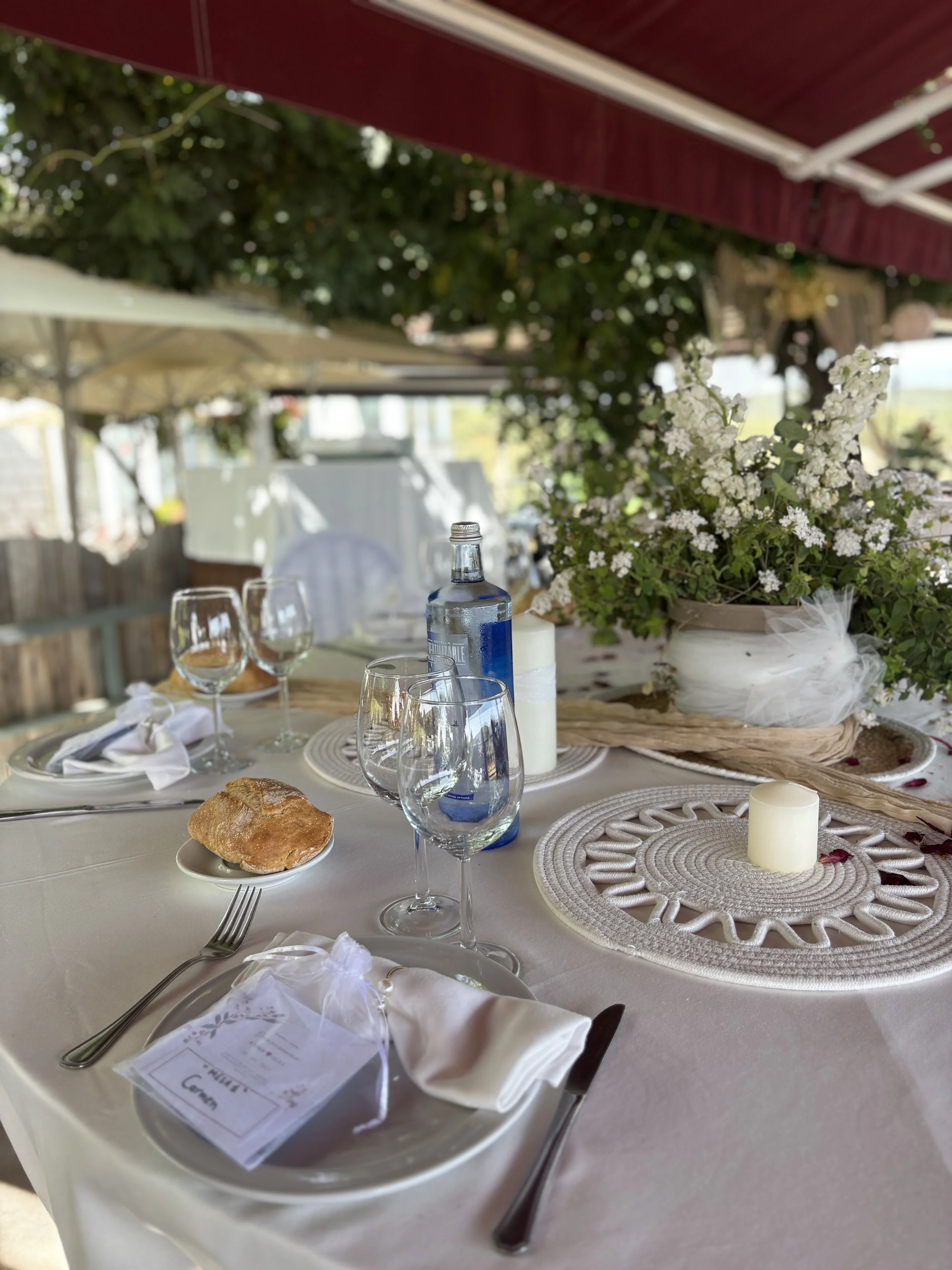 Mesa decorada con un florero con flores blancas, platos, copas y un pan en un evento al aire libre con sombrillas y árboles.
