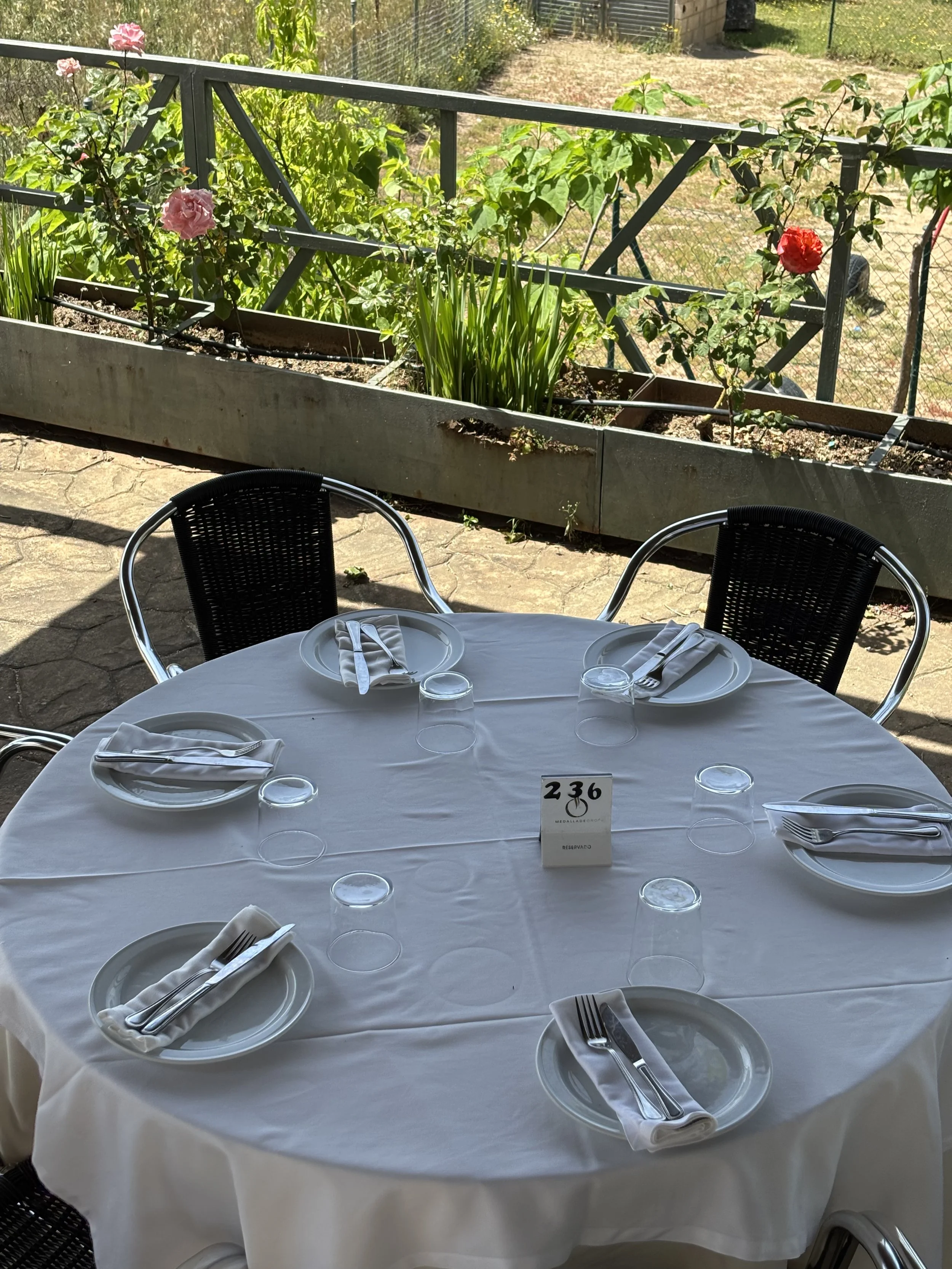 Mesa redonda con manteles blancos y utensilios de cena, rodeada de sillas, en un espacio al aire libre. En el centro de la mesa hay un cartel con el número 236 y la palabra 'Reservado'. En el fondo, un jardín con plantas y flores, y una cerca metálica.