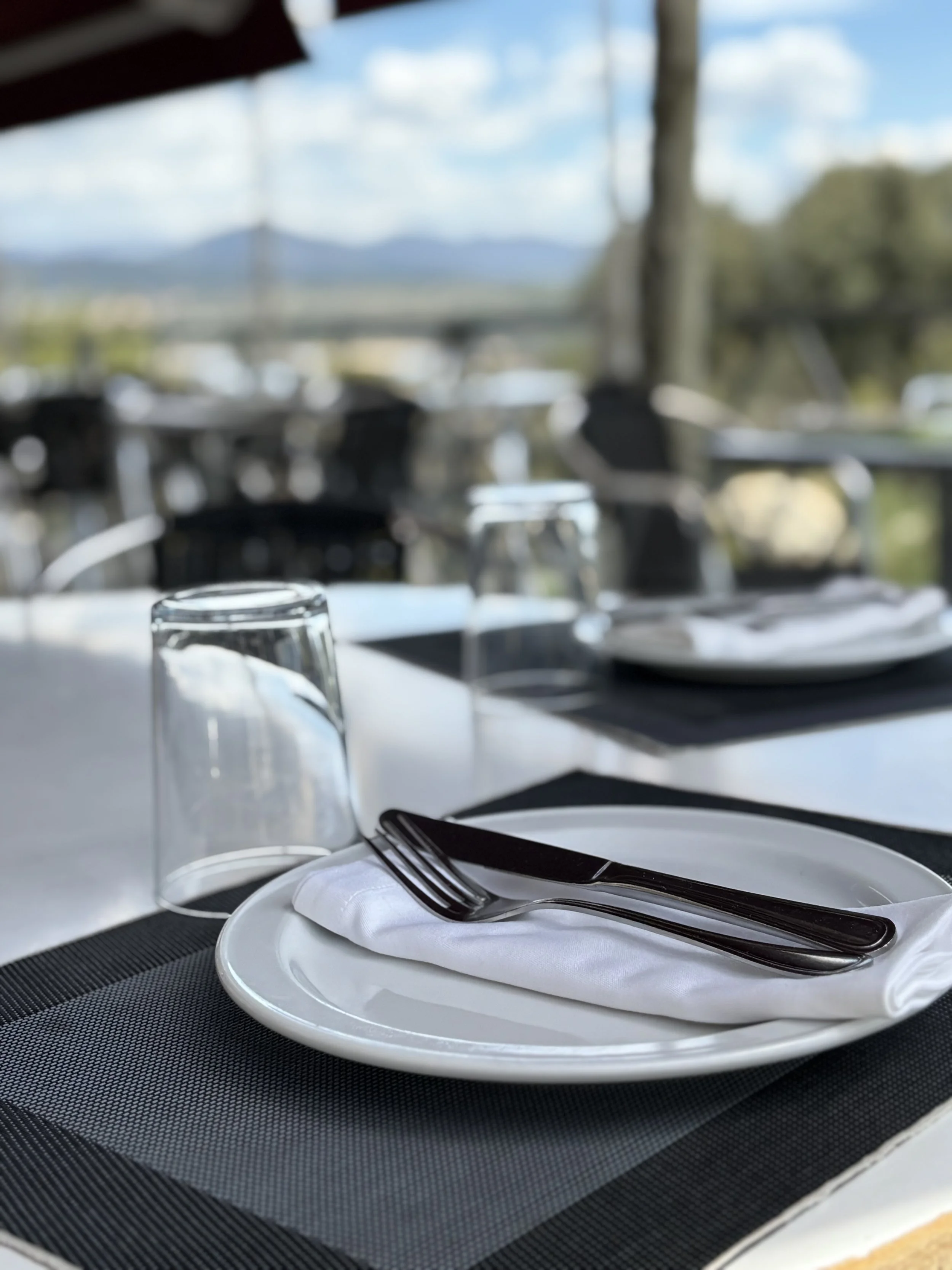 Mesa puesta para comer con platos, vasos y cubiertos sobre un mantel negro en un ambiente con vista al aire libre y montañas.