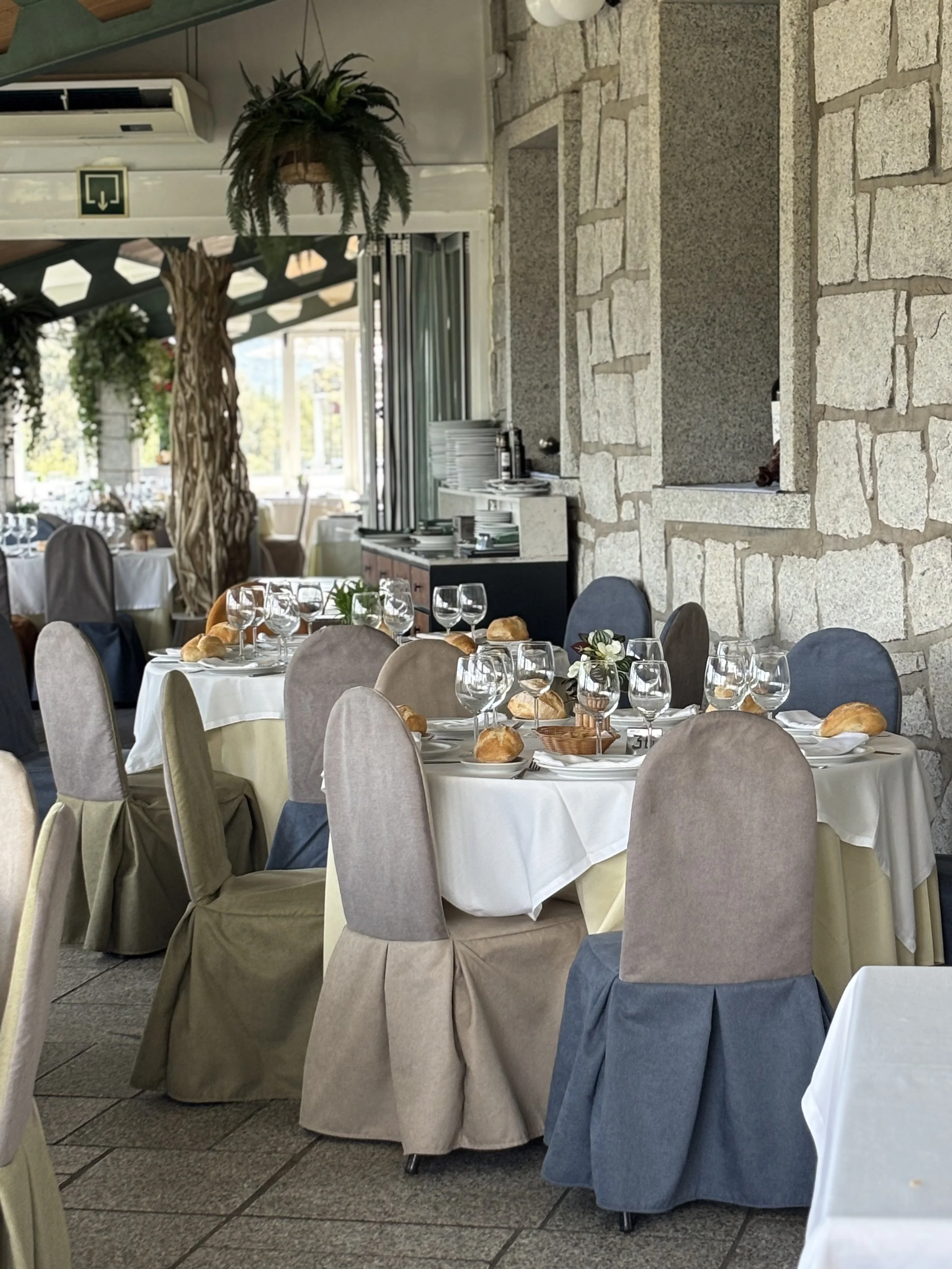 Mesas decoradas para una comida en un restaurante, con manteles blancos, cubiertos, copas y panes, en un ambiente interior con paredes de piedra y ventanas grandes.