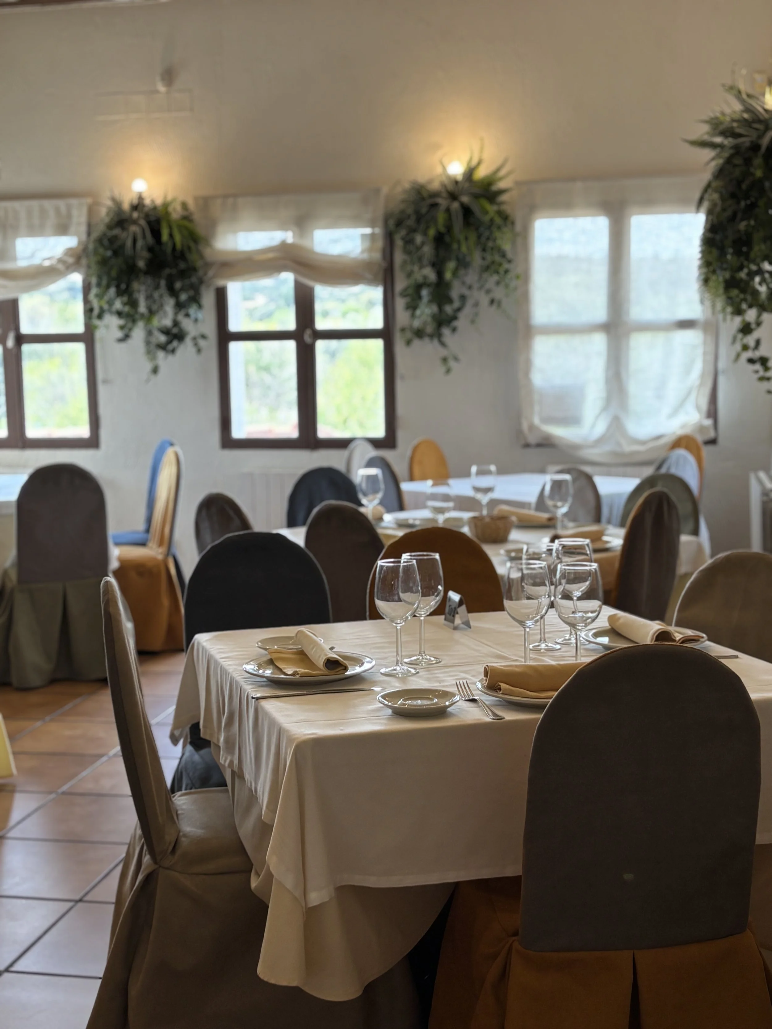 Habitación decorada para un evento con mesas vestidas y copas en un ambiente bien iluminado con ventanas y cortinas.