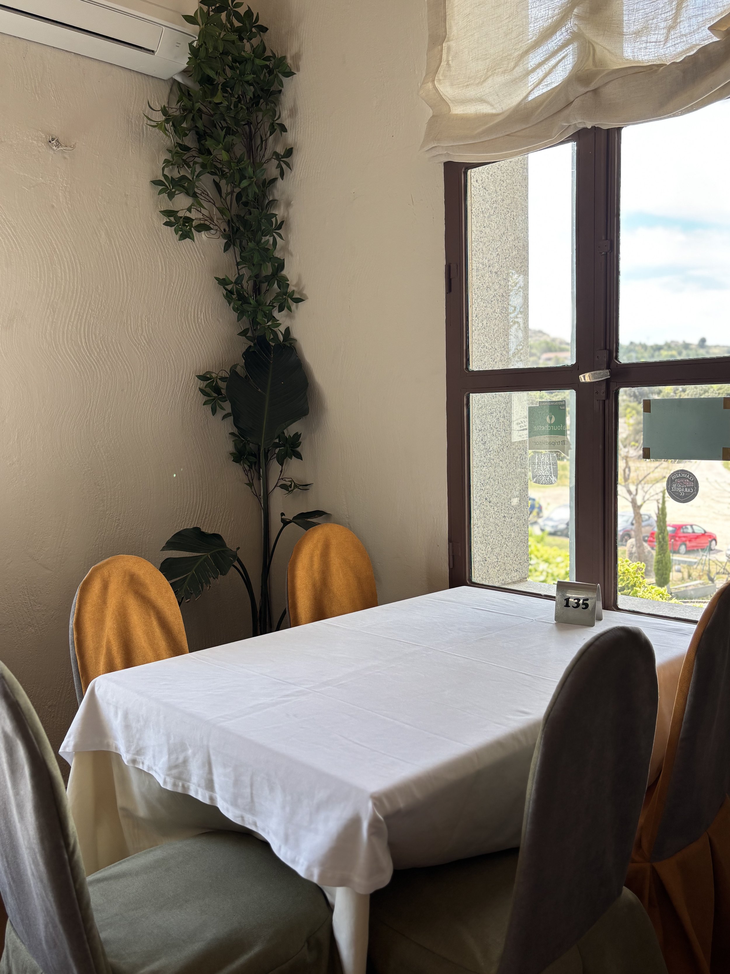 Mesa de comedor cubierta con una tela blanca, rodeada de sillas con fundas de diferentes colores, con una planta decorativa en la esquina y una ventana grande que muestra un paisaje exterior con coches y árboles.