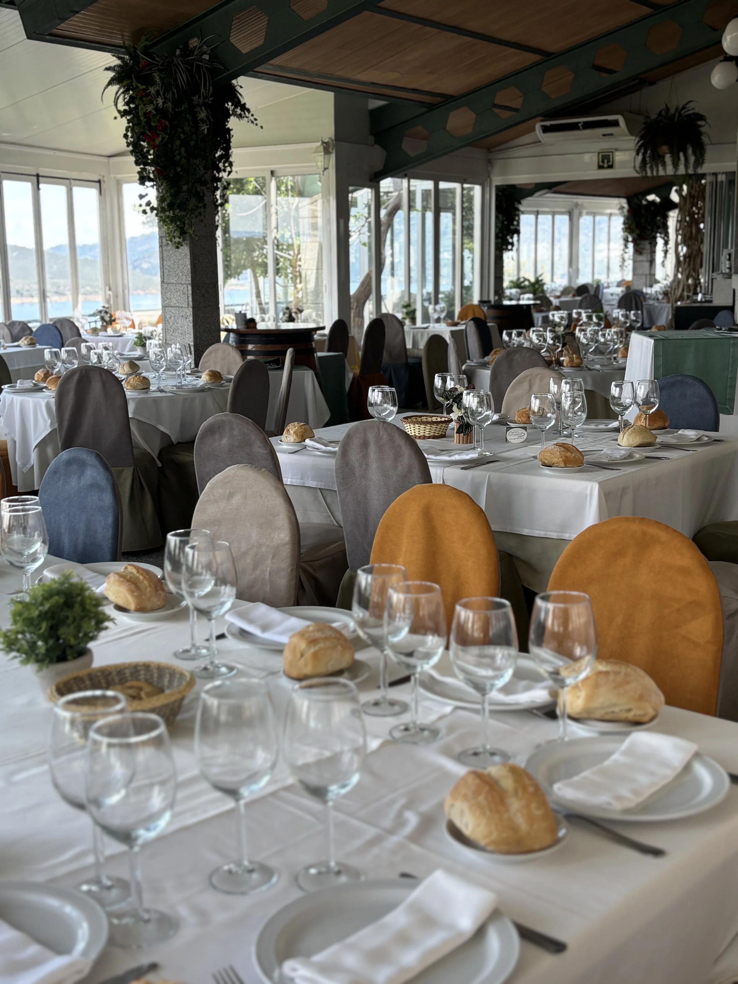 Una sala de eventos decorada con mesas cubiertas con manteles blancos y servilletas dobladas, con copas de vino y pan en cada lugar, en un ambiente con grandes ventanas que ofrecen vista al exterior con árboles y un paisaje montañoso.
