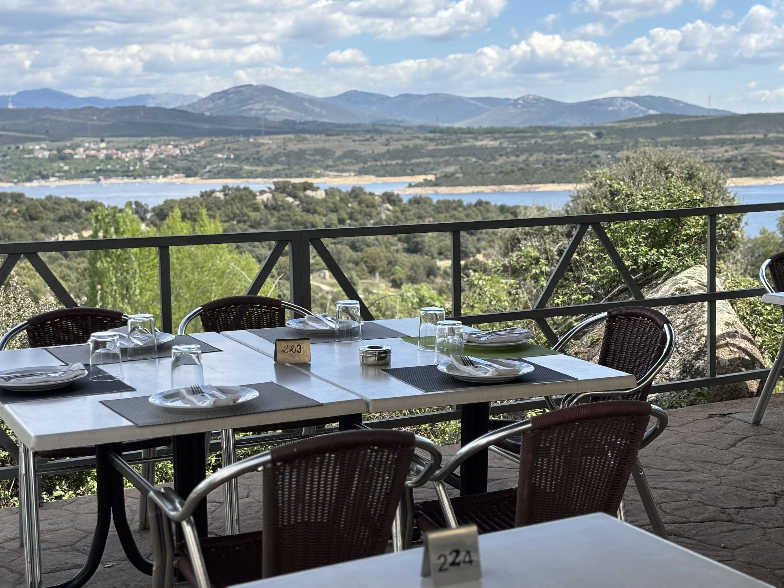 Mesa de comedor en un restaurante al aire libre con vista a un paisaje con montañas y un río, mesas con platos, vasos y cubiertos, y un ambiente tranquilo y natural.