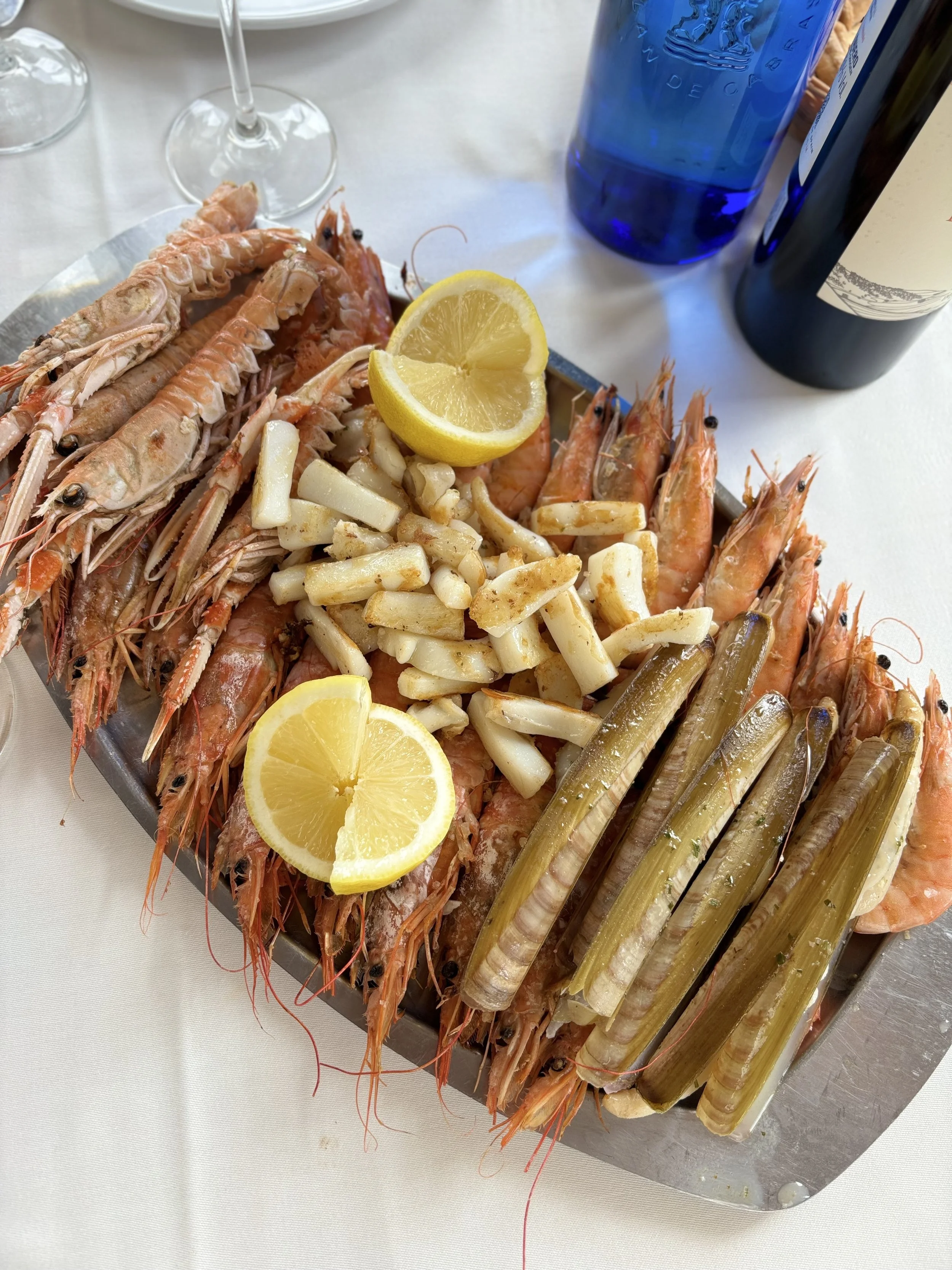 Plato con mariscos variados, limón, pasta, y verduras en un plato de metal, acompañado de copas y botellas sobre una mesa