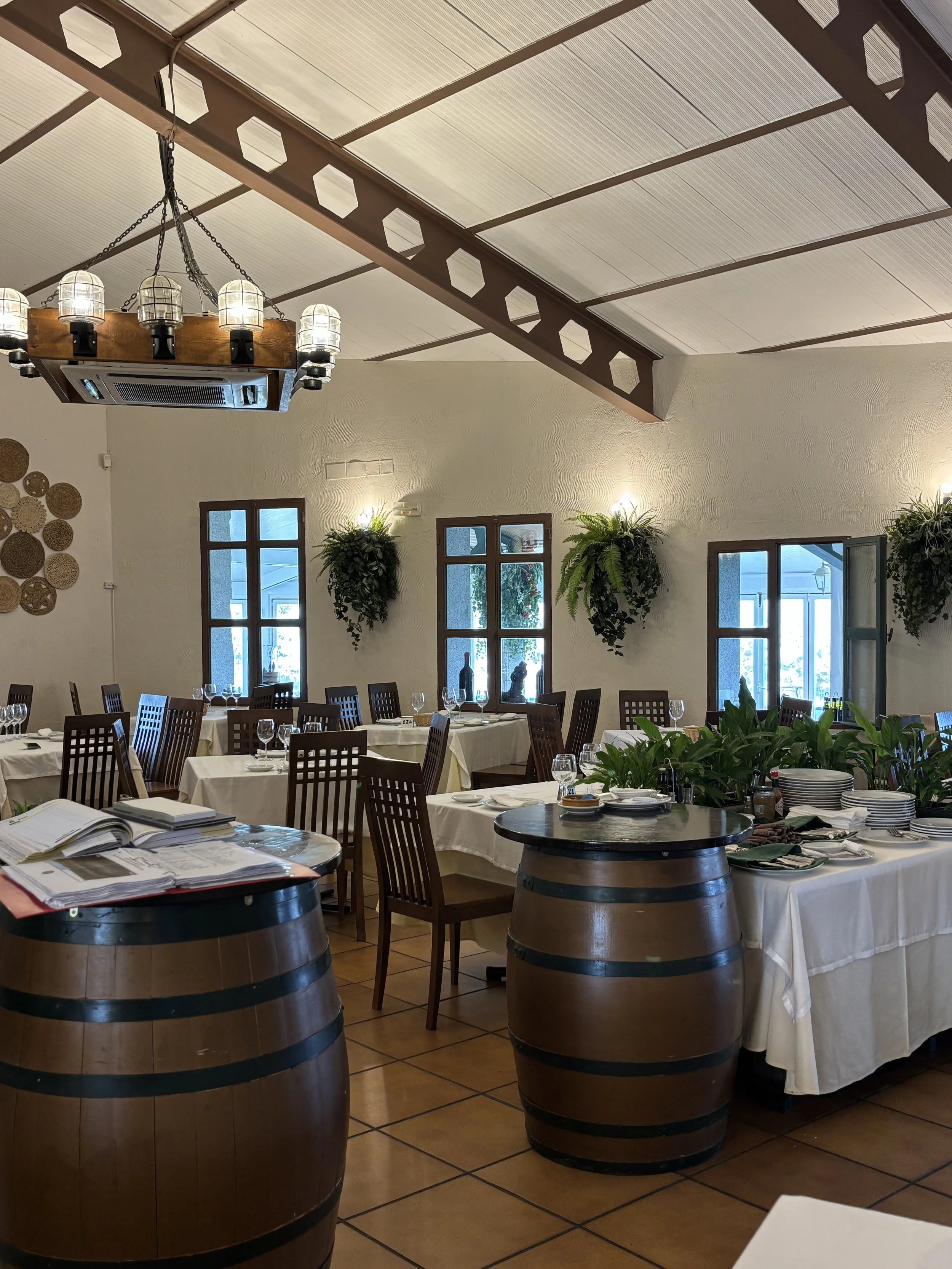 Vista interior de un restaurante con mesas vestidas con manteles blancos, sillas de madera y decoración con plantas y cerámicas en las paredes. Hay una lámpara de techo de estilo rústico y ventanas con marcos de madera que dejan entrar luz natural.