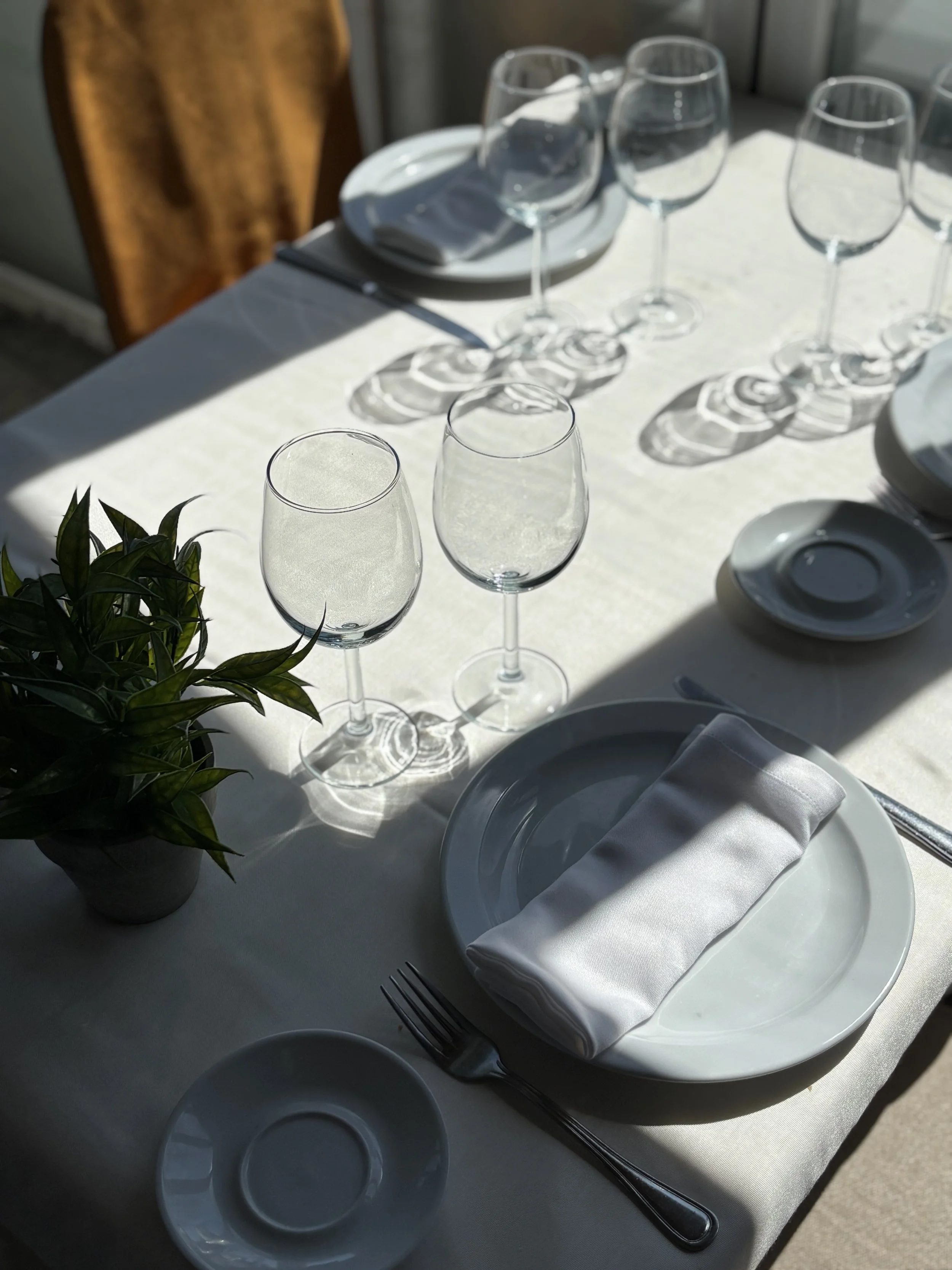 Mesa puesta con copas de vino, platos, utensilios y un florero con plantas verdes en un ambiente bien iluminado.