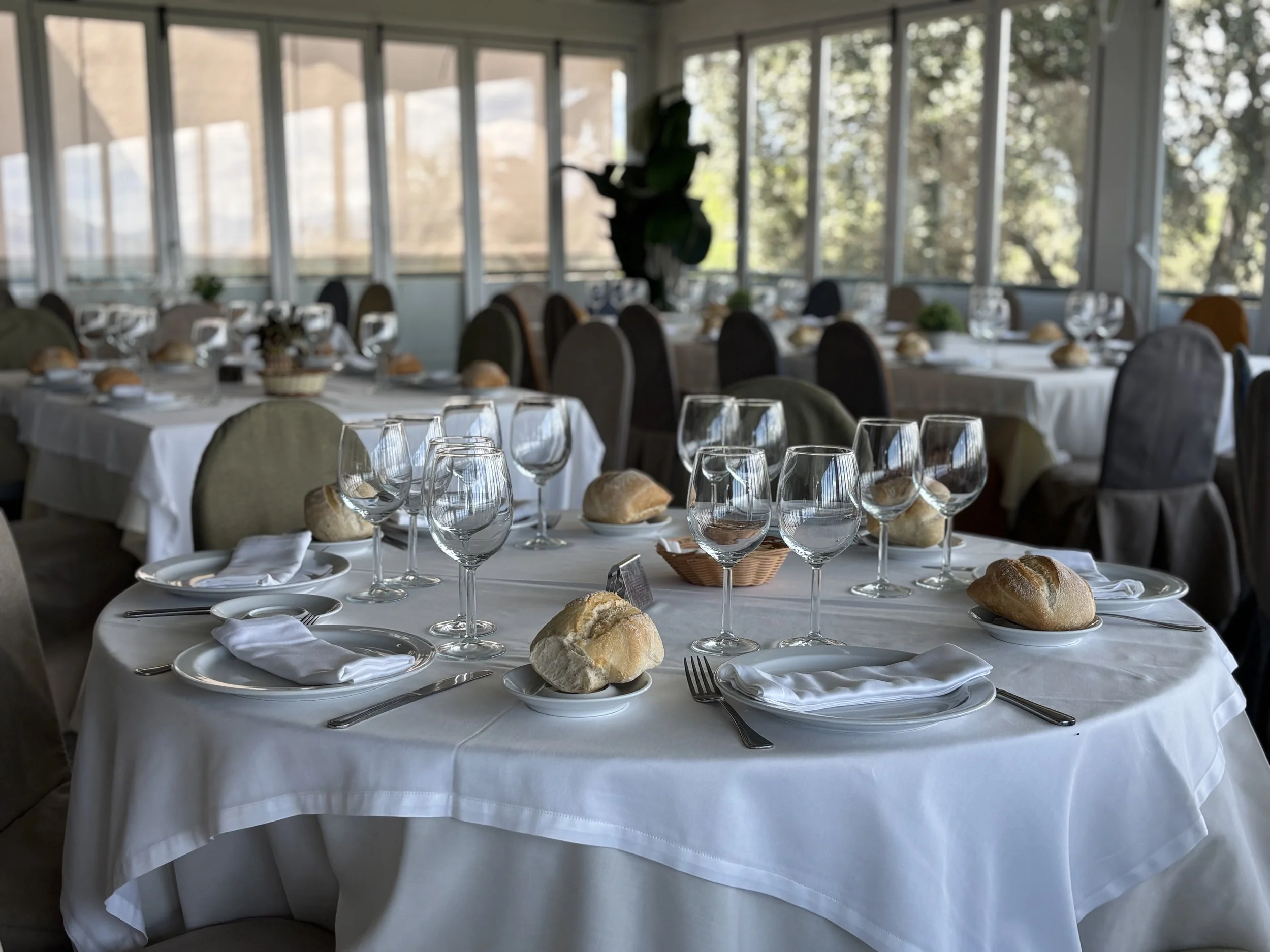 Mesas preparadas para una comida o evento, con manteles blancos, vasos, platos, pan y cubiertos en un espacio con ventanas grandes que dejan entrar luz natural.