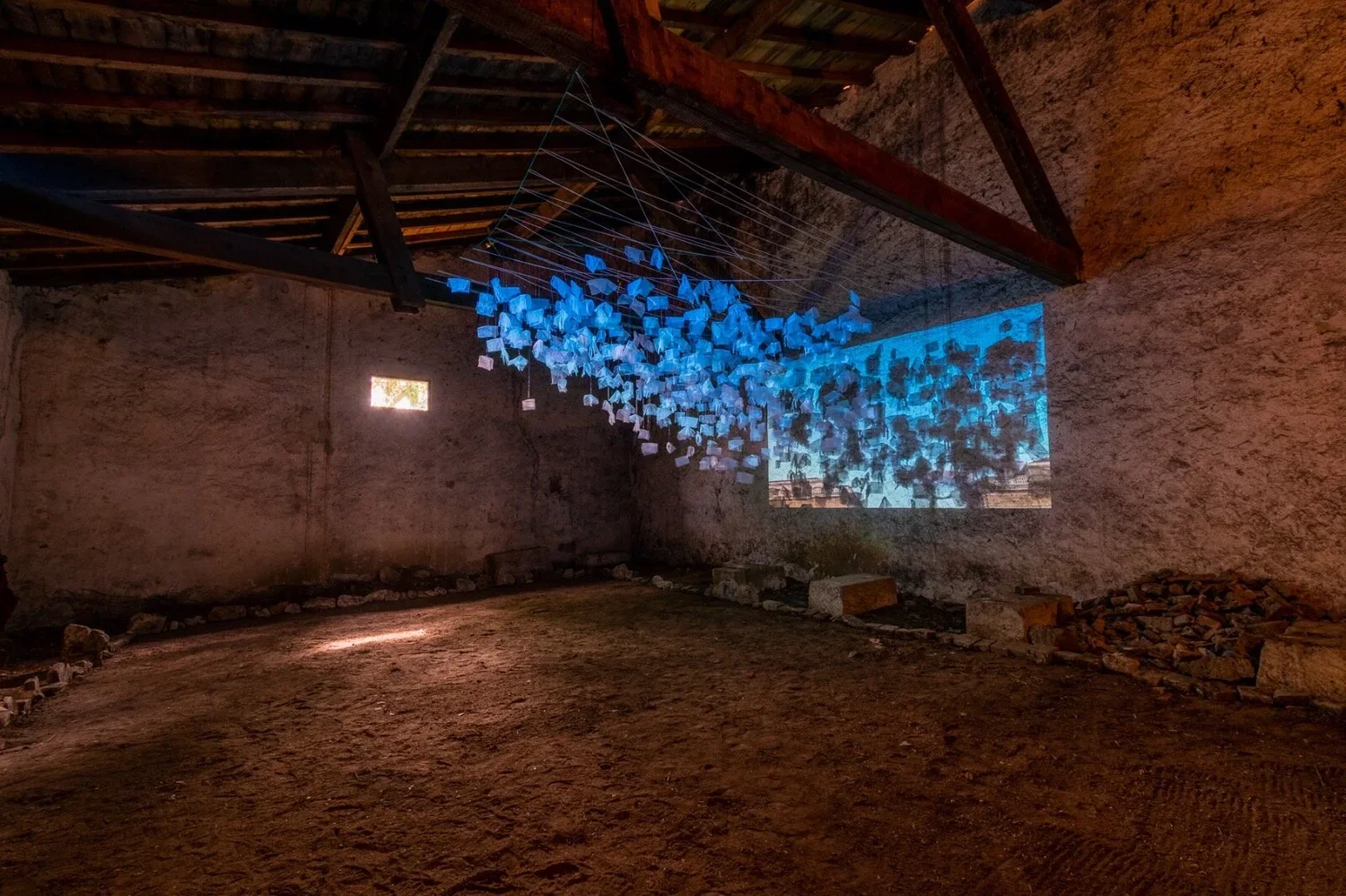 An art installation inside a rustic barn with stone walls and wooden roof beams. It features a suspended sculpture made of multiple blue and white geometric shapes connected by thin strings. A digital projection on the wall displays an abstract patte