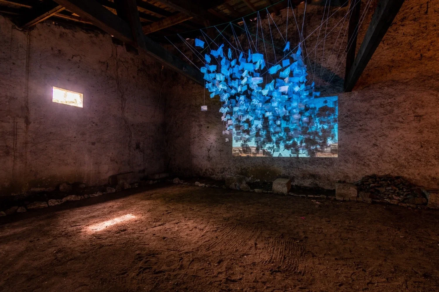 An art installation inside a rustic, unfinished room with dirt floor and stone wall, featuring a suspended sculpture of many blue paper or fabric pieces hanging from strings, and a video projection on the wall.