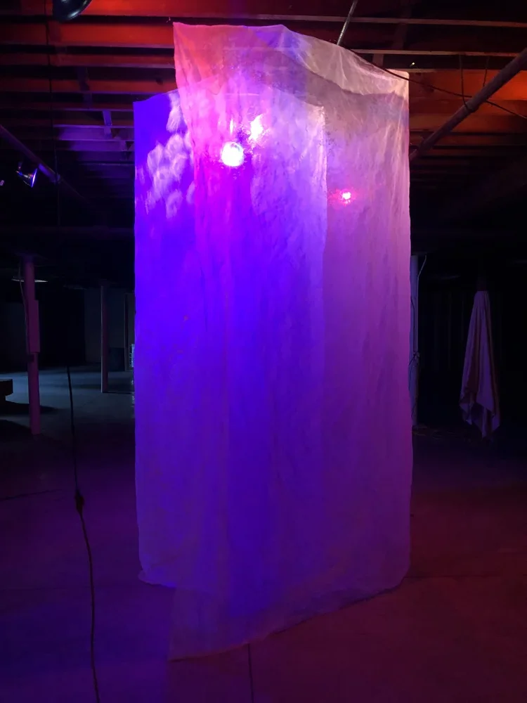 A large illuminated fabric structure illuminated by purple, blue, and pink lights, hanging in a dark indoor space with a visible ceiling and some furniture.