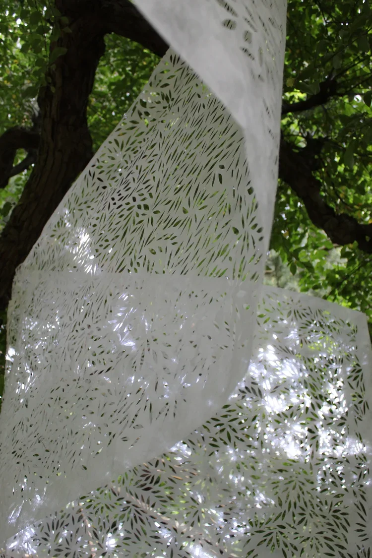 Close-up of white decorative fabric with leaf patterns, hanging from a tree in a natural outdoor setting with green leaves and sunlight filtering through.