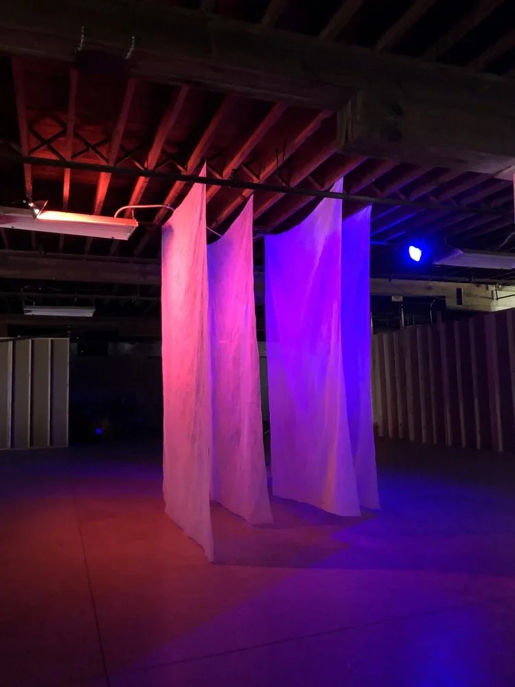 Three colorful fabric curtains hanging from the ceiling in a dimly lit space with purple and pink lighting.