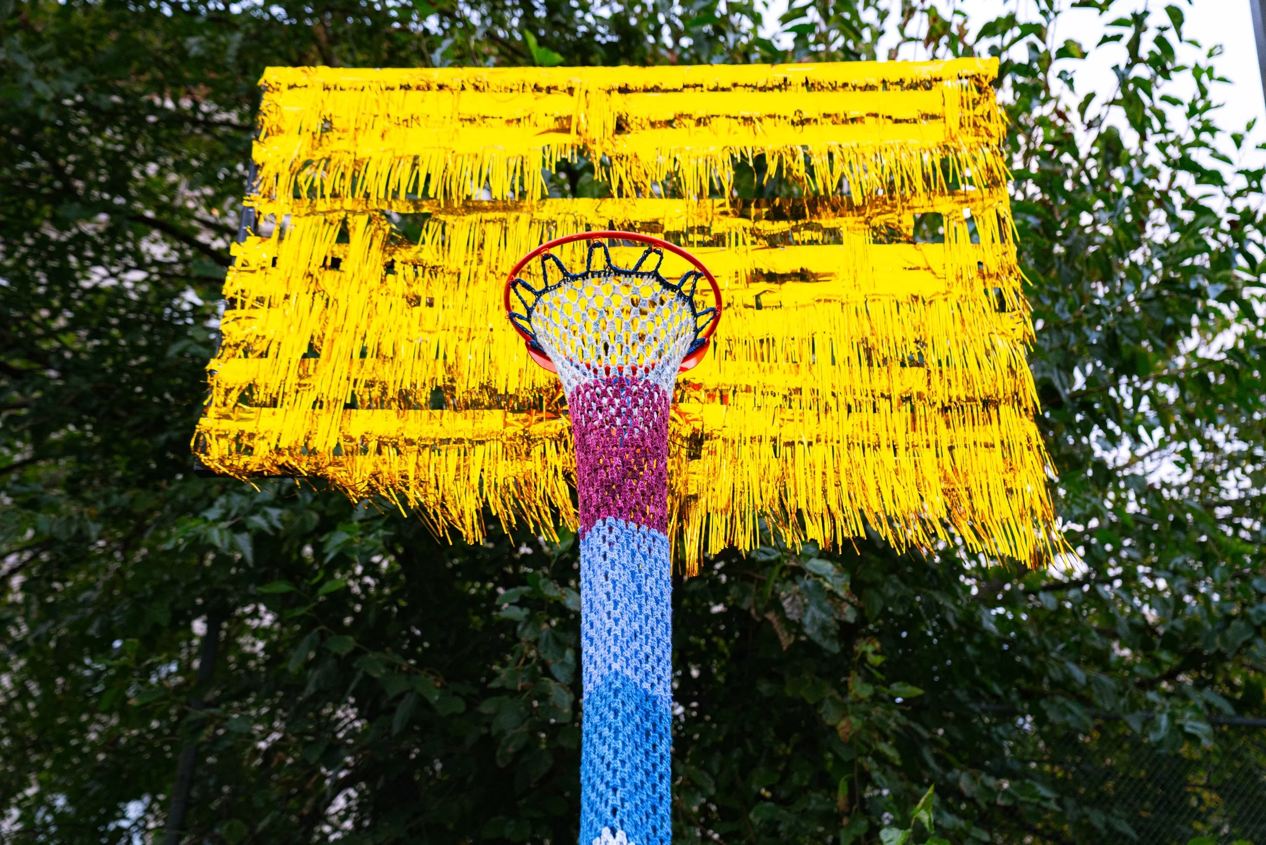 Colorful basketball hoop with a yellow fringed backboard and a multicolored net, viewed from below against a background of green trees.