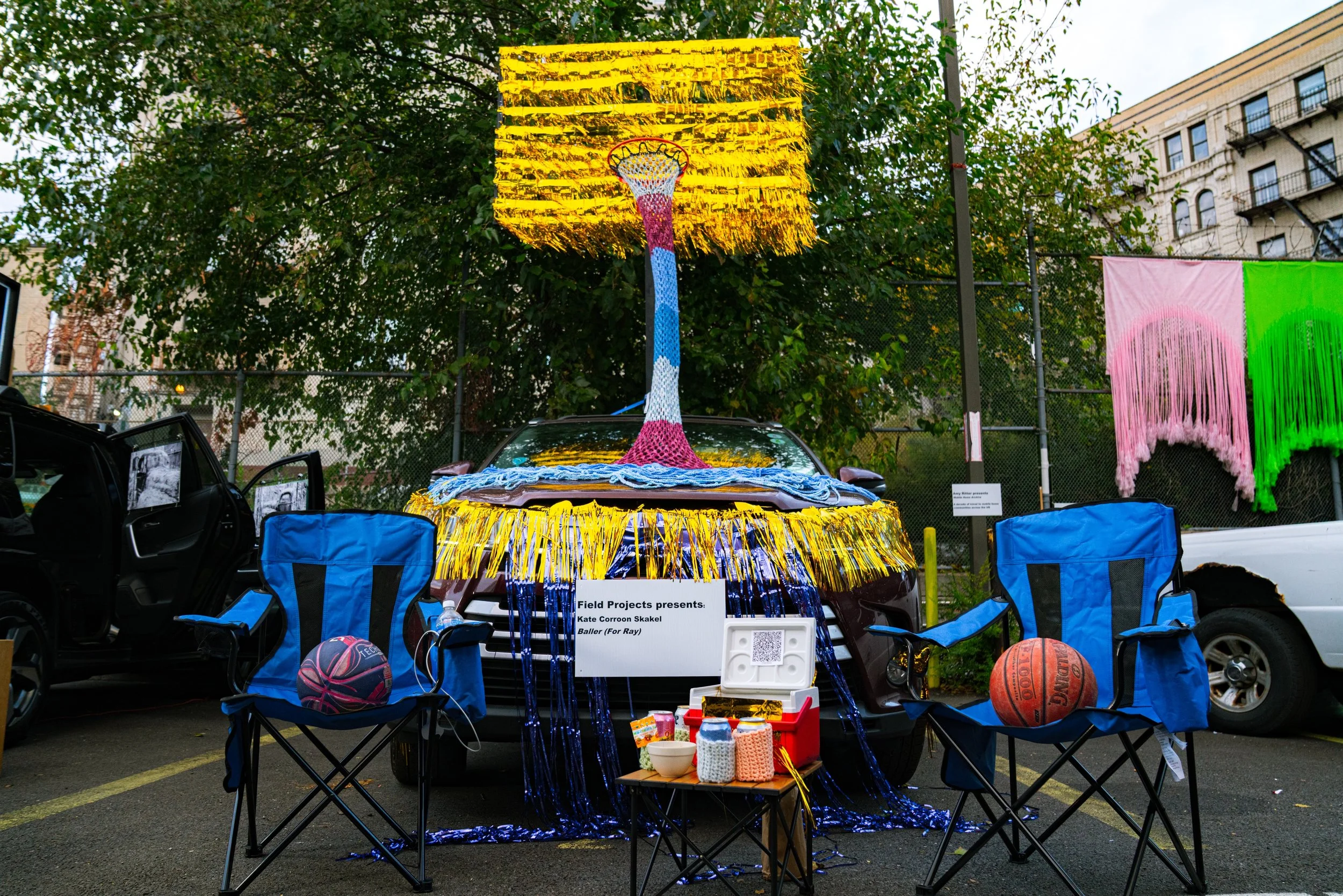 Decorative basketball-themed art display on a car and adjacent chairs with basketballs, featuring a large hanging basketball hoop sculpture surrounded by yellow and blue fringe, set up in a parking lot with trees and buildings in the background.