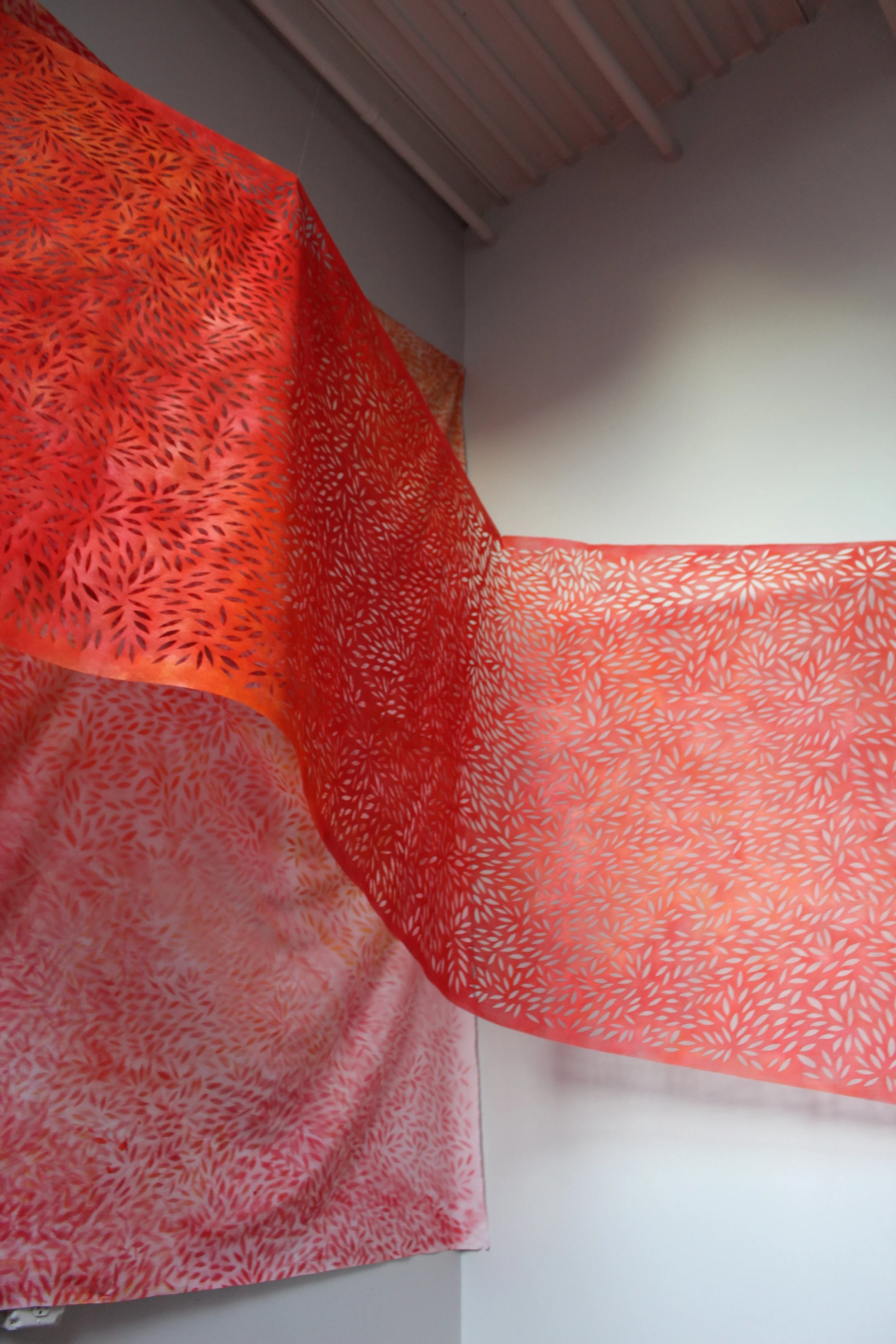 Red fabric with intricate white leaf patterns hangs from a ceiling with a metal roof, partially draped over a white wall.