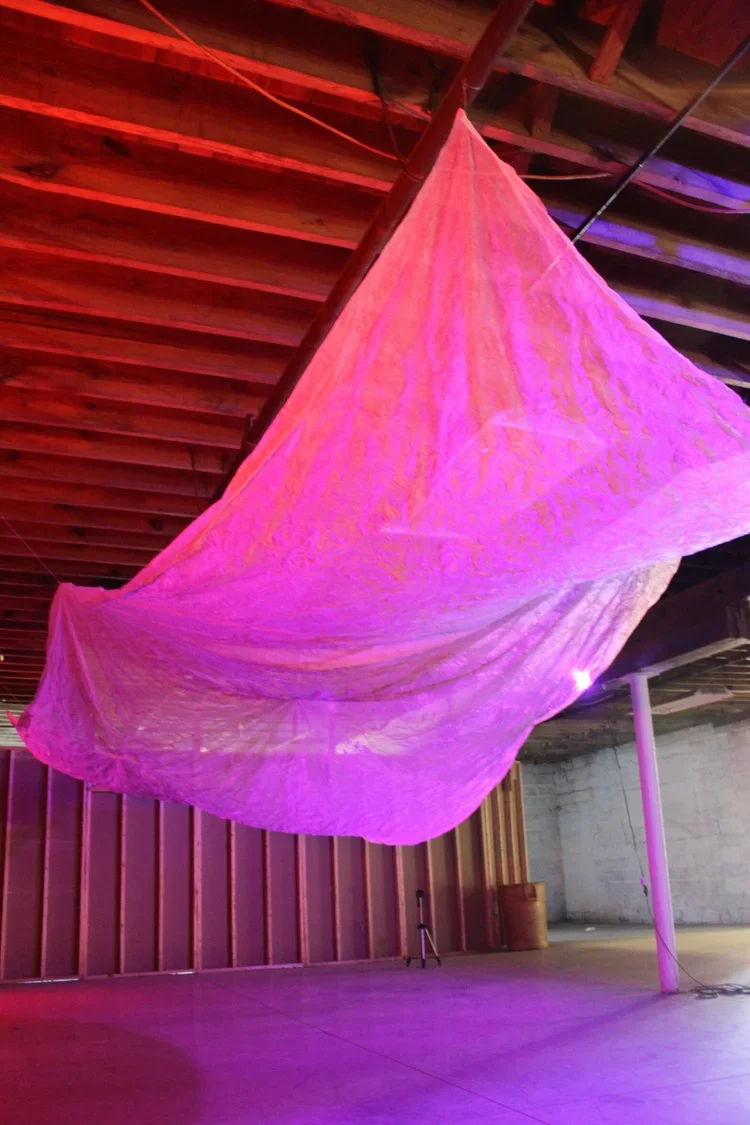 Large, pink, fabric kite hanging from a wooden ceiling in a warehouse or studio space, illuminated by purple lighting.