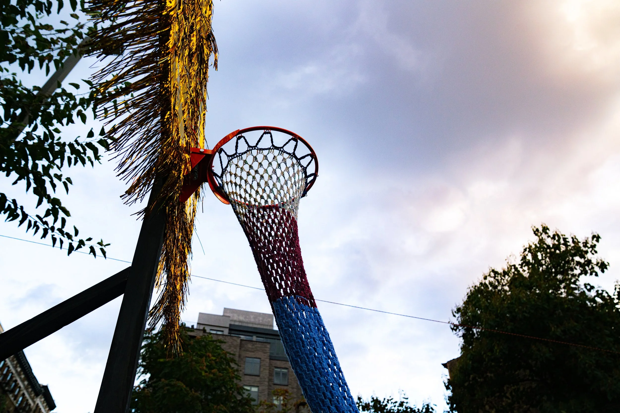 Basketball hoop with a net resembling a colorful party decoration, set outdoors with trees and a building in the background, under a partly cloudy sky.