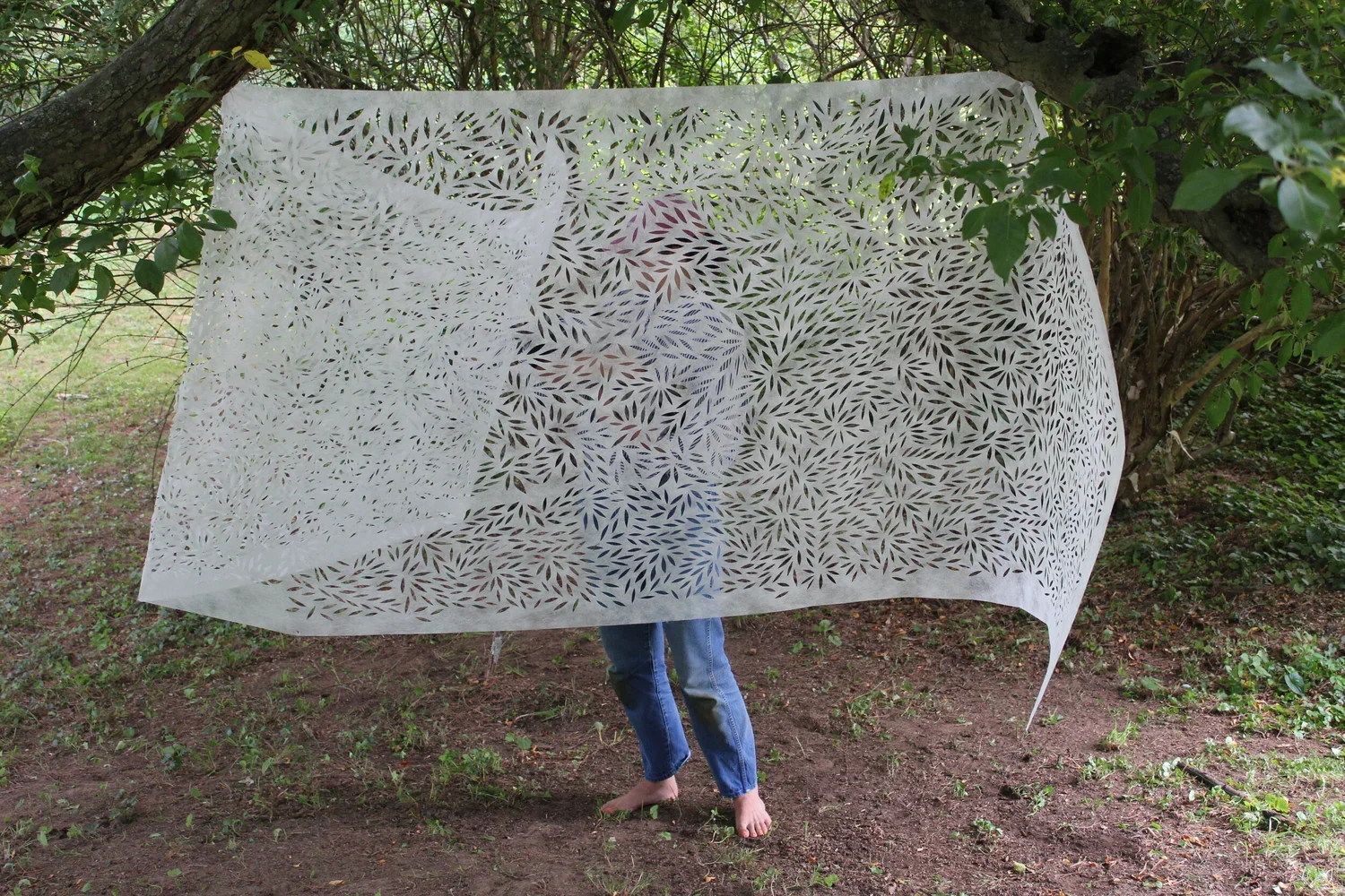 Person standing outdoors behind a patterned white fabric hanging from tree branches, with only their legs visible.